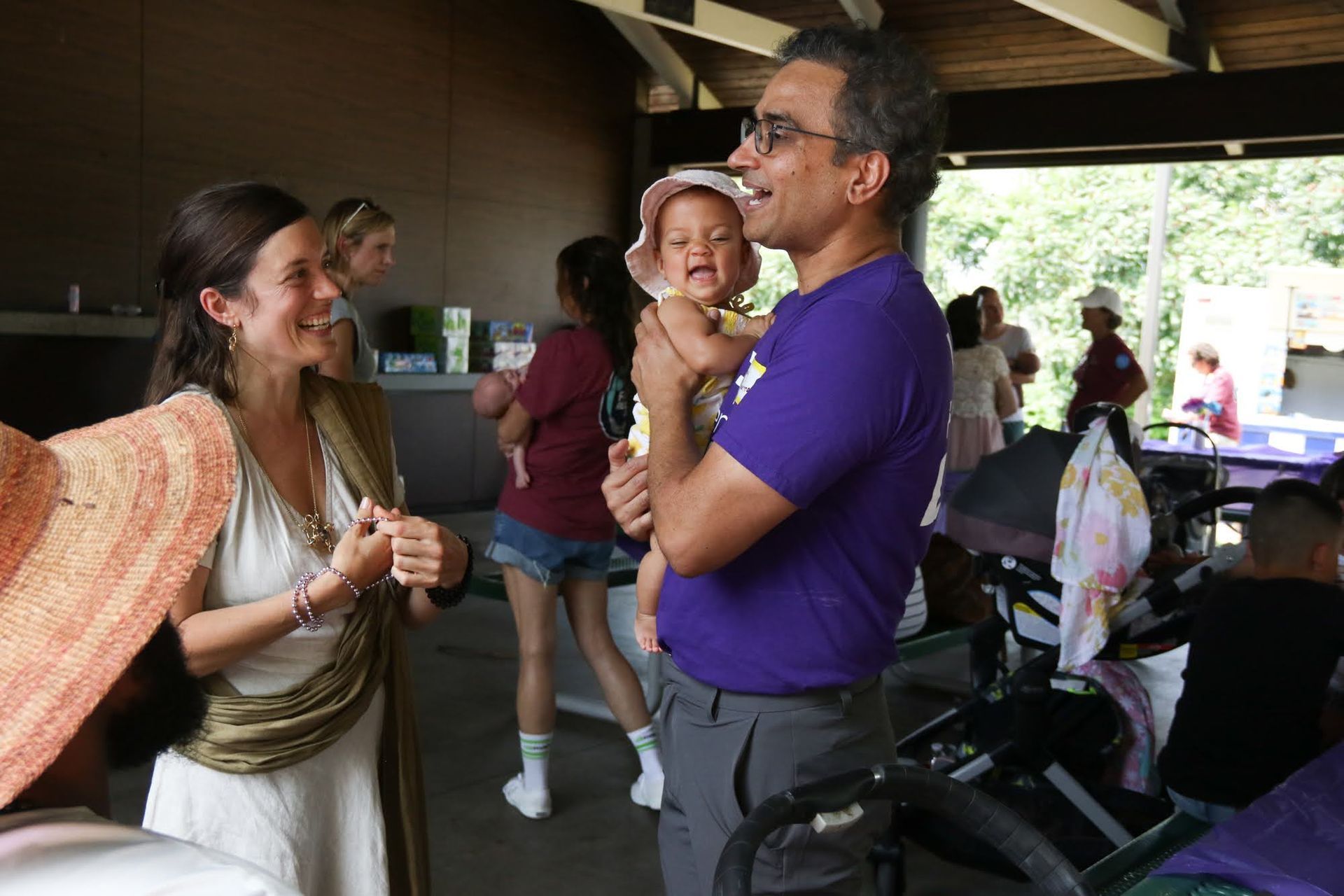 Woman talks with man holding a baby; they are outside near a park shelter with others around.