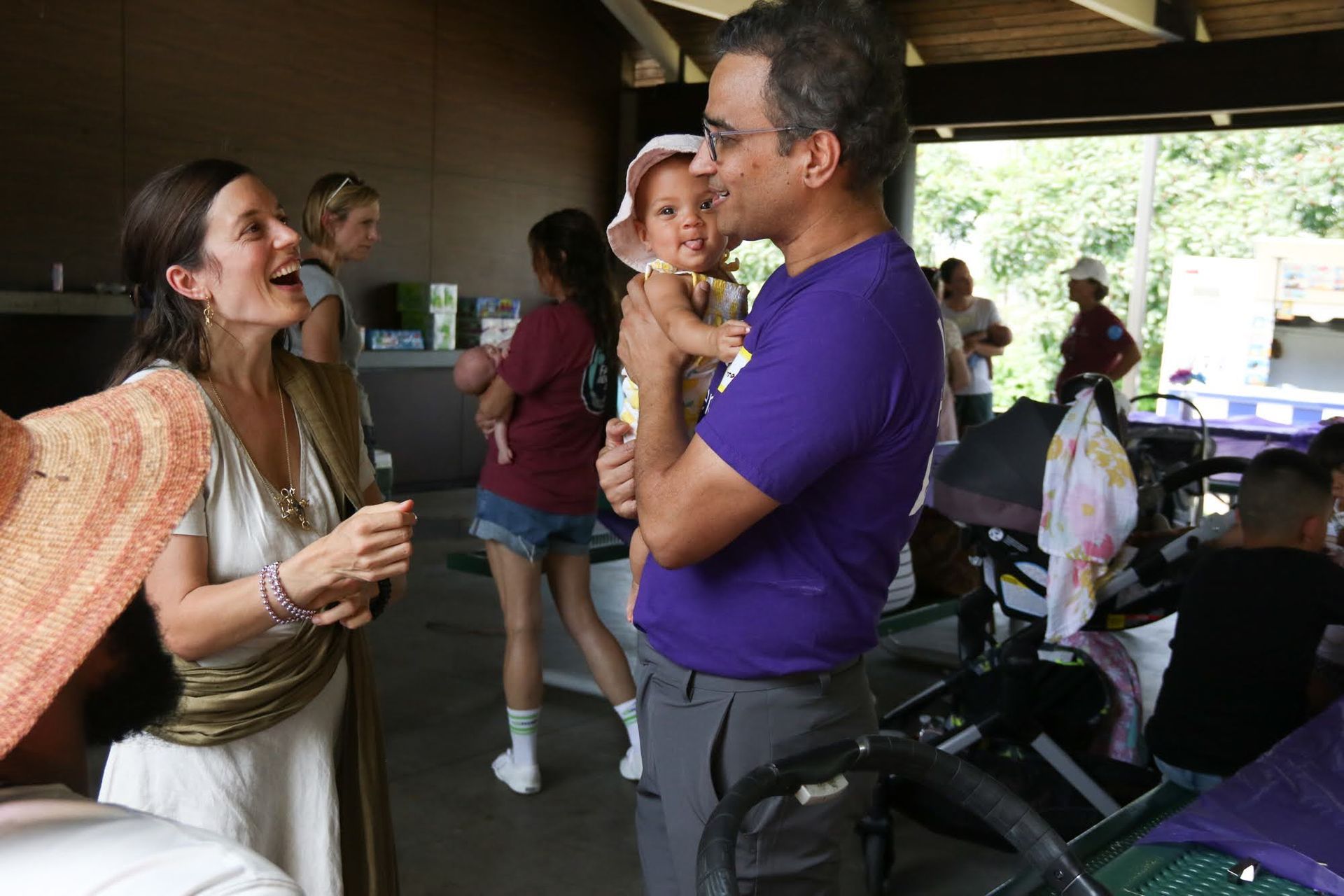 A man holds a baby; a woman smiles and talks to them outdoors, near a park shelter with others.