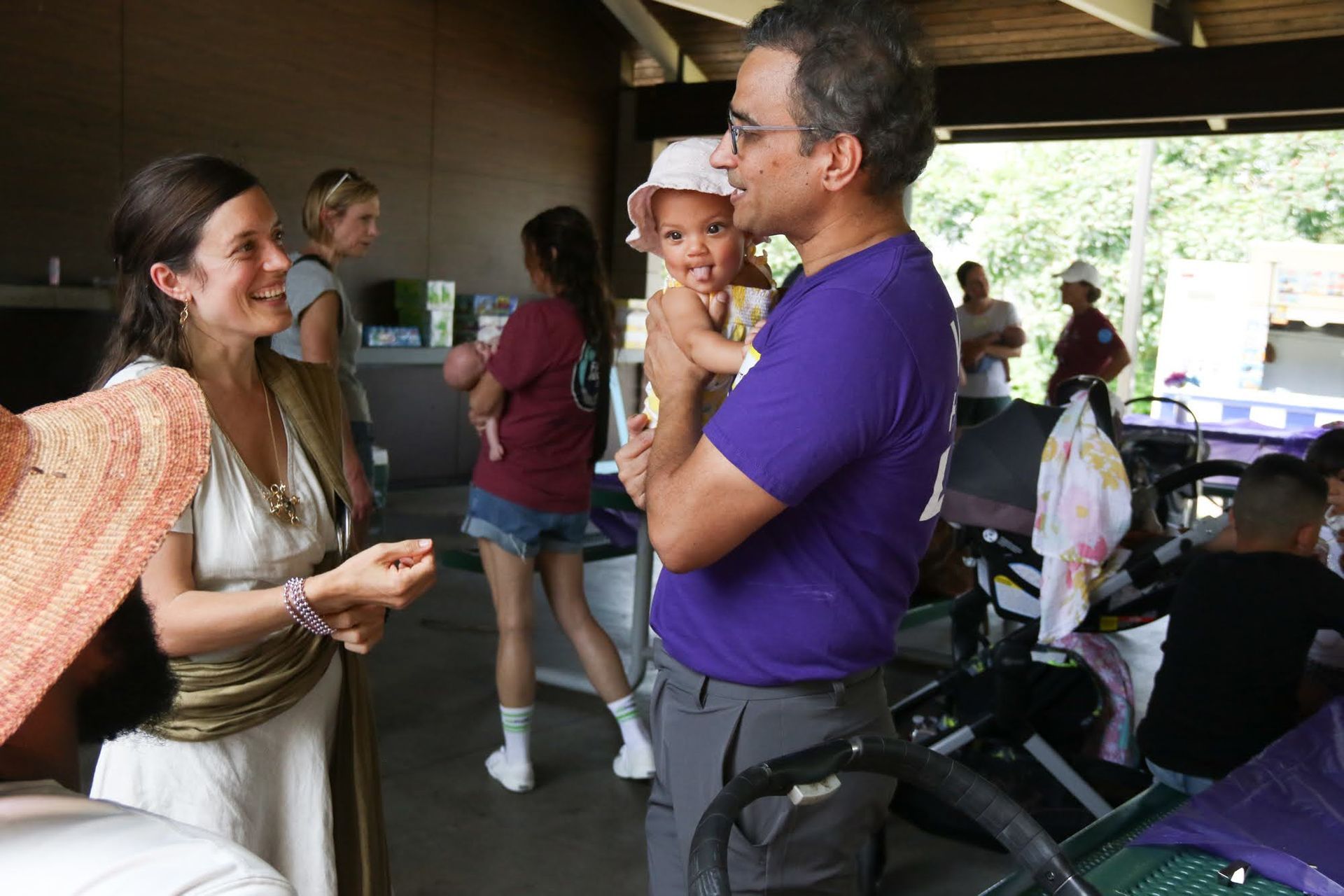 People at an outdoor gathering, a woman with a baby, a man in purple holding a baby, socializing under a shelter.