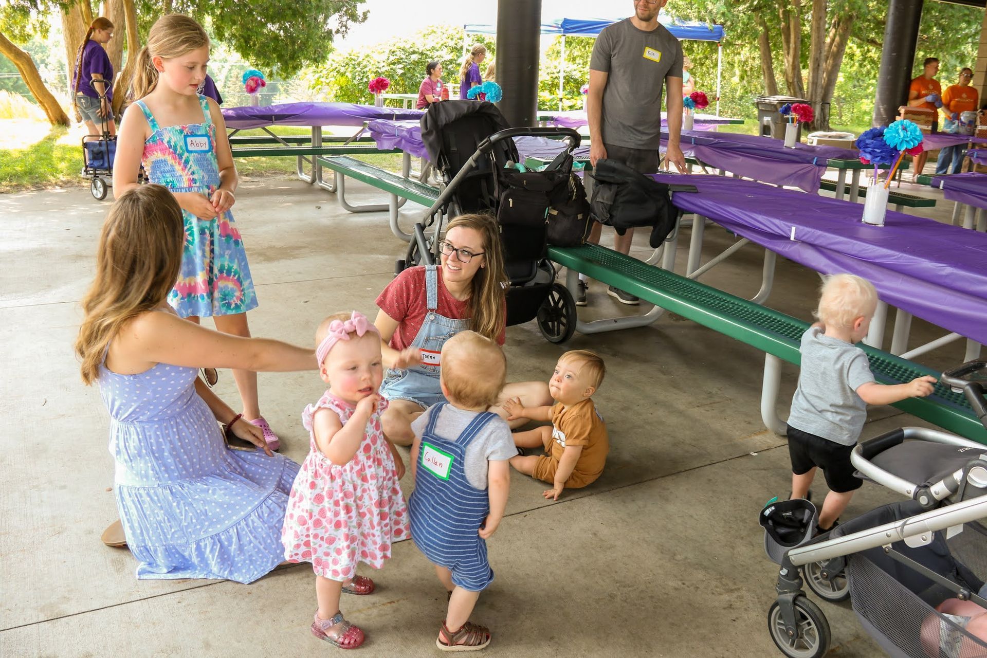 People at a gathering in a pavilion. Children playing near adults, picnic tables set up.