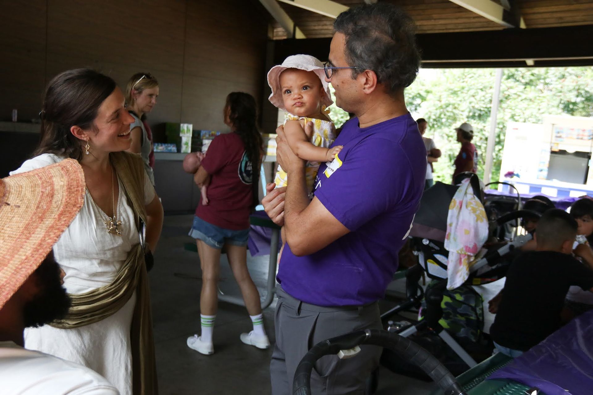 Man holding baby wearing sun hat; woman in white dress smiles, outdoor gathering.