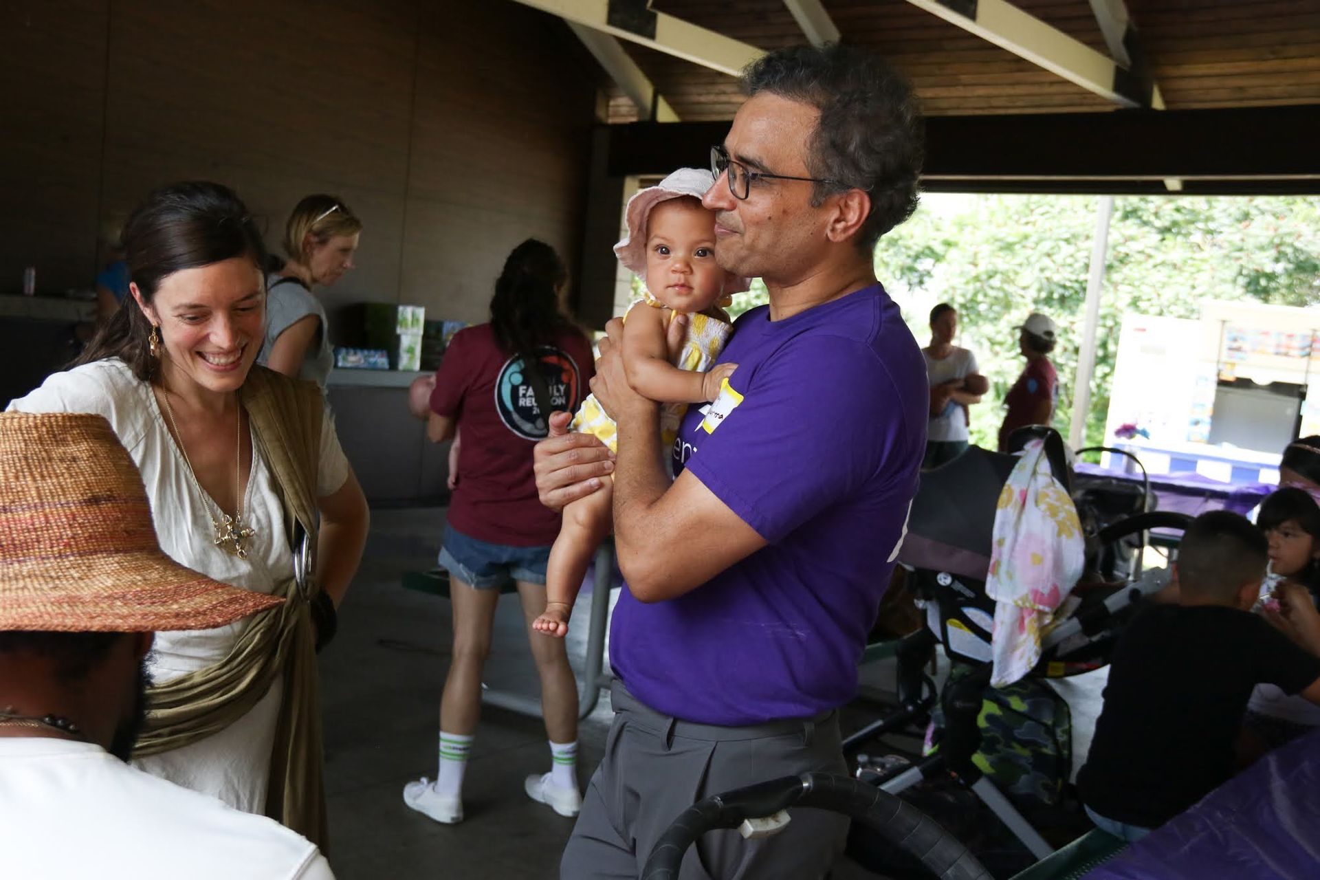 Man holding baby, woman smiling nearby, at an outdoor gathering. Other people in the background.