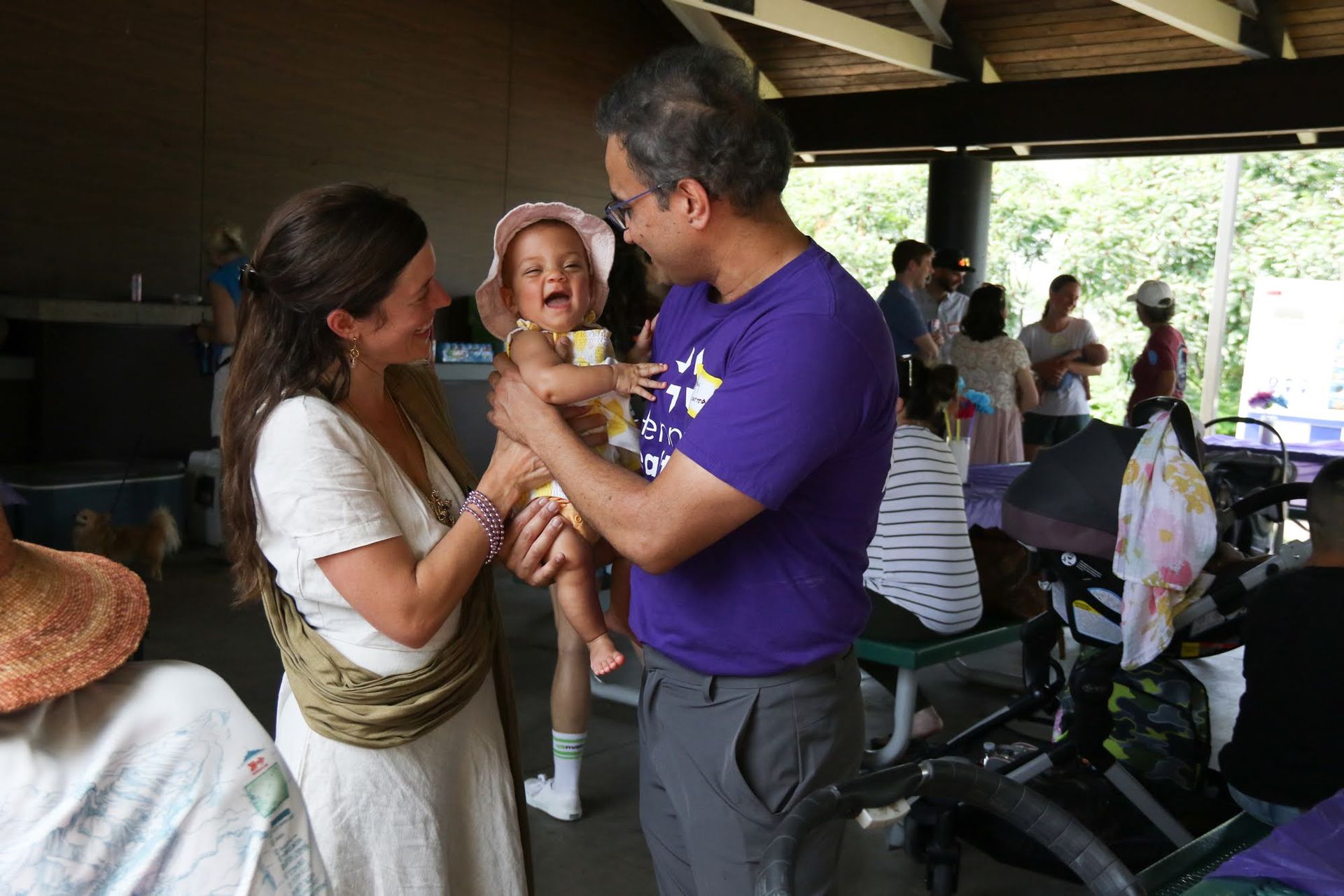 Parents holding a baby laughing at a party in a park pavilion.