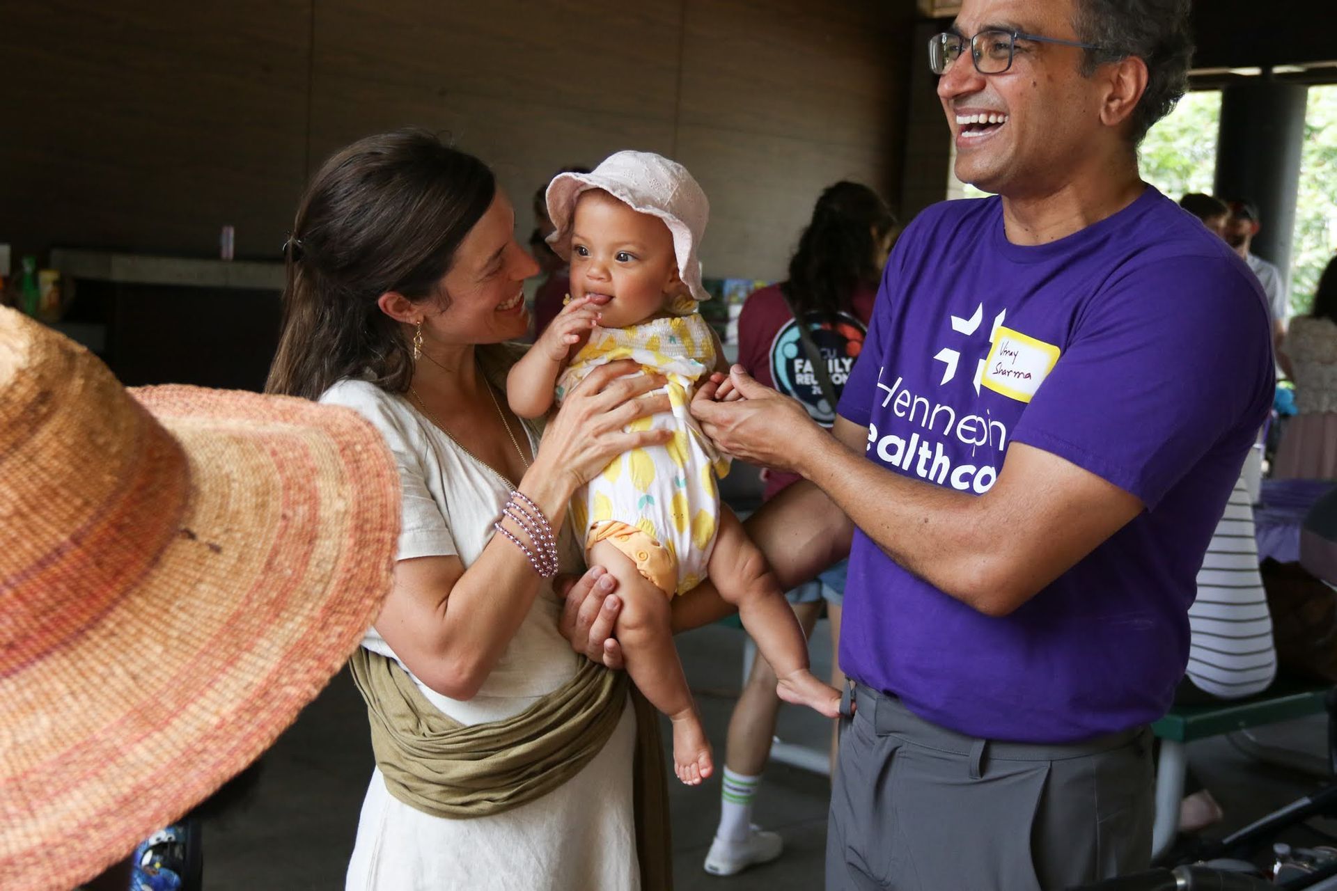 Woman holds baby, man smiles. Outdoor gathering, sunlight.
