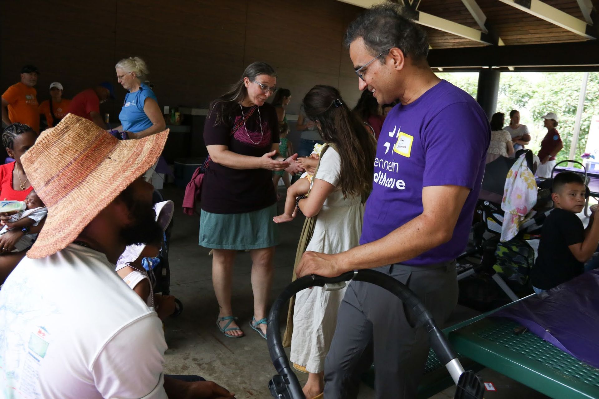 People at a park shelter. One man holds a baby. Another woman gestures. Others sit at tables.