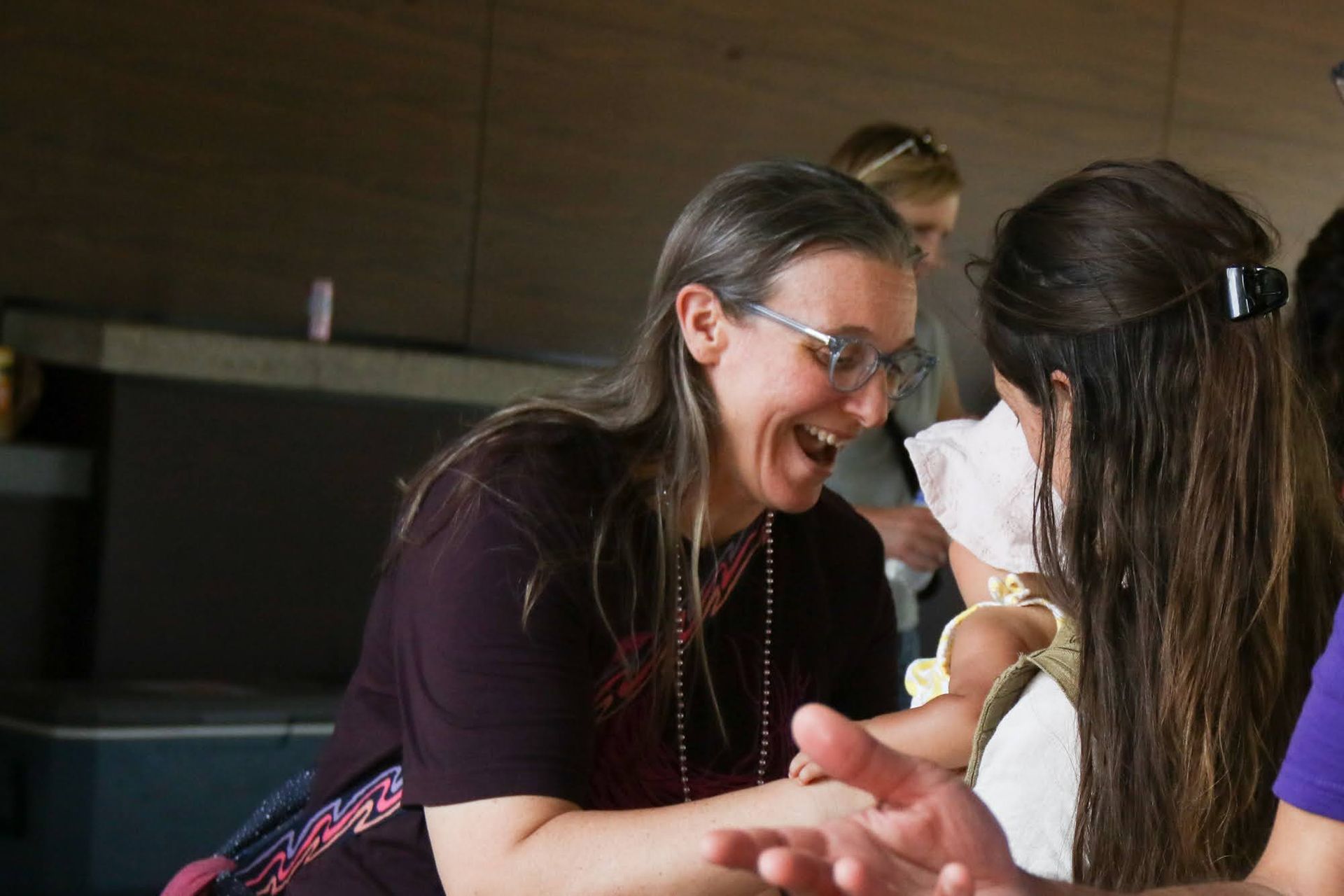 Woman laughs while interacting with someone holding a baby. Indoors, natural light.