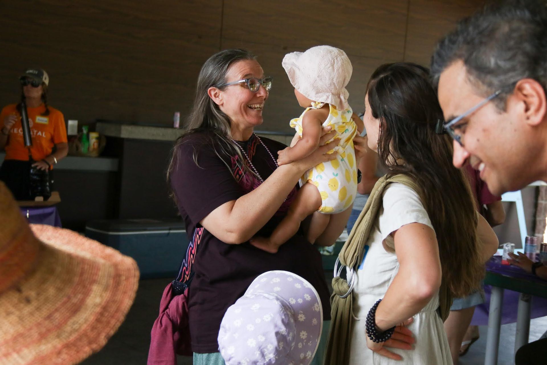 Woman holding a baby smiles at another woman; others look on indoors.