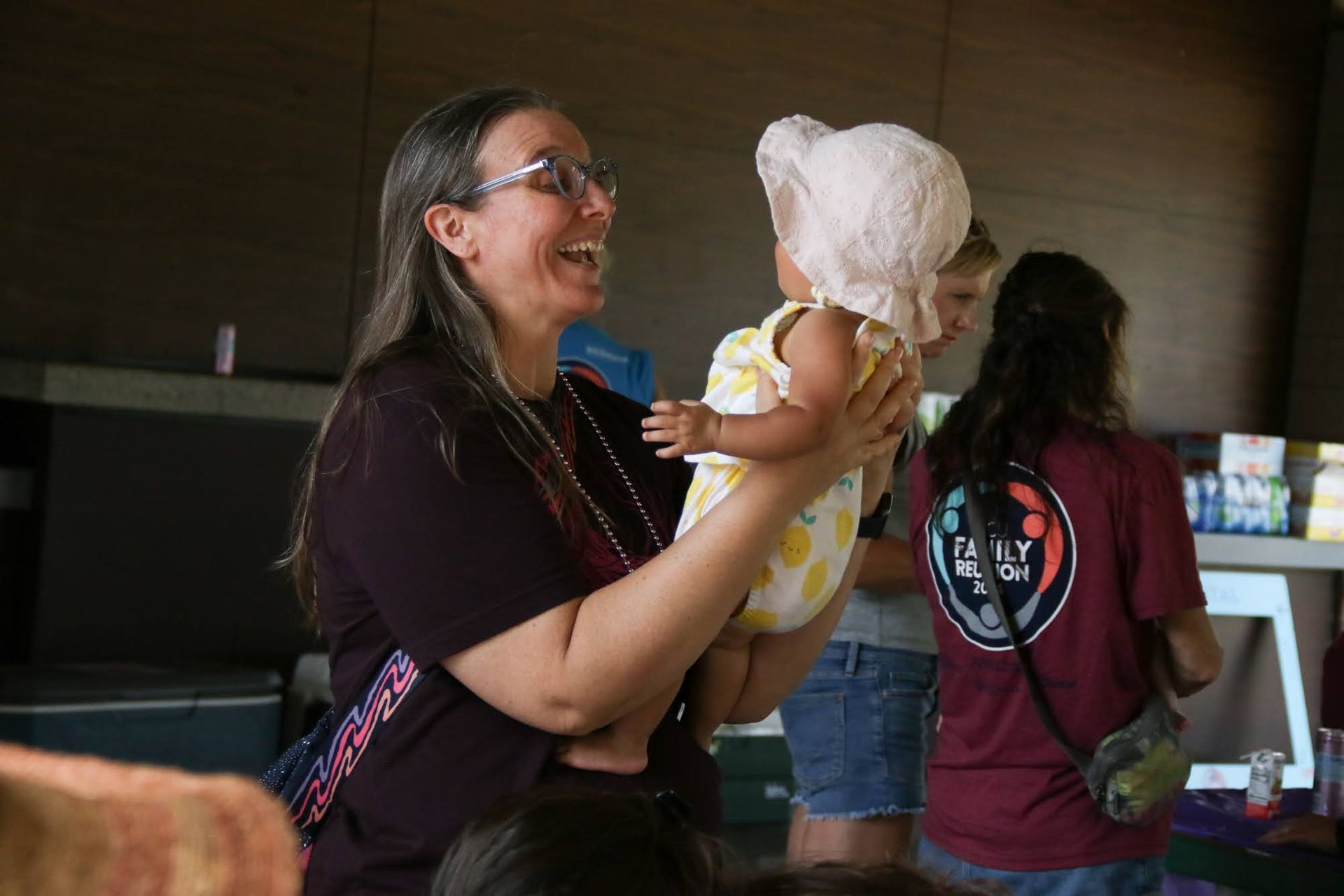 Woman laughs, holding a baby wearing a sun hat and yellow onesie. Others stand nearby indoors.