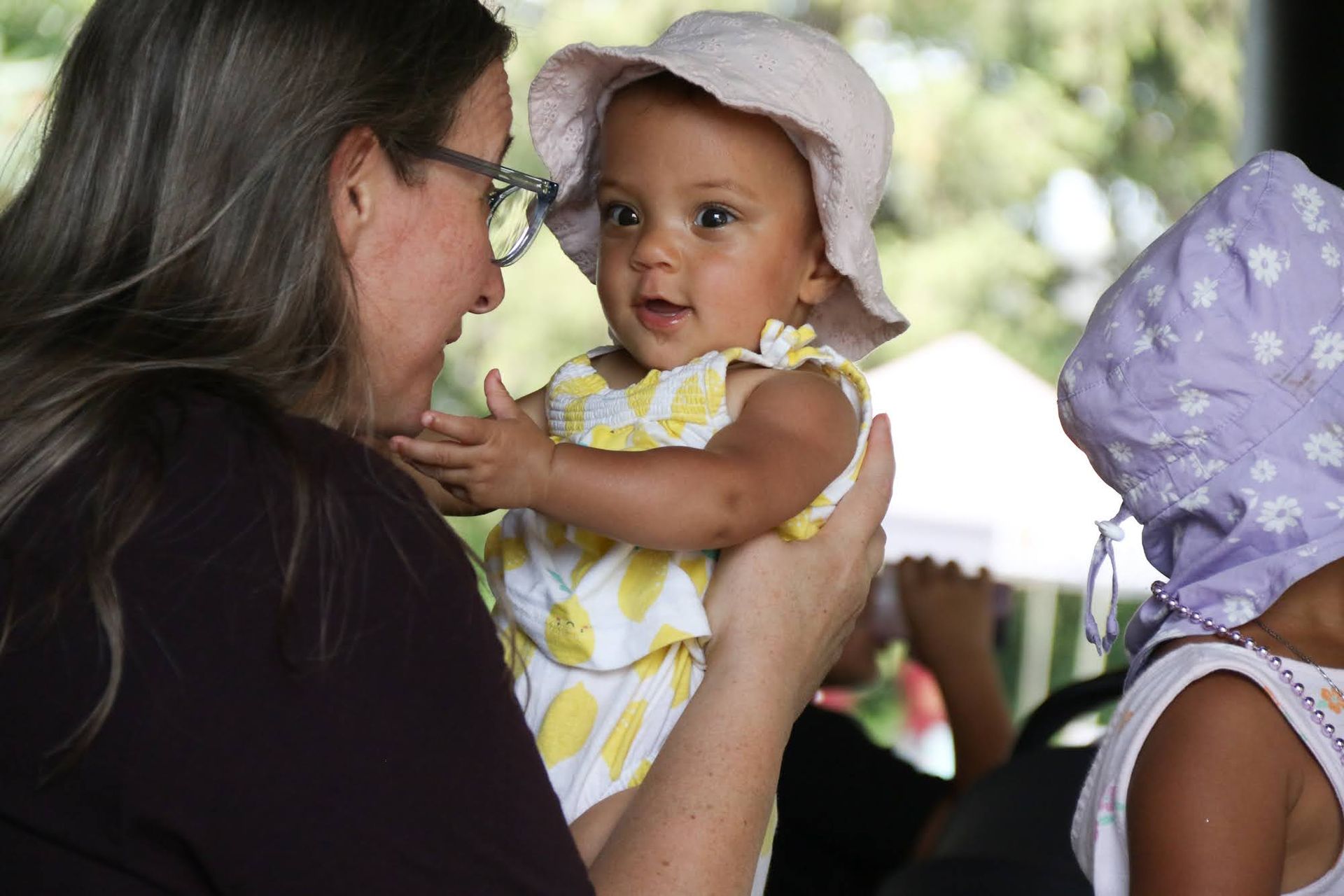 Woman holding a baby wearing a sunhat and yellow outfit, outdoors.