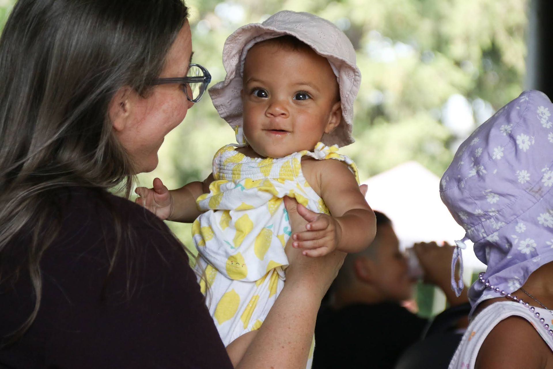 Woman holding up a smiling baby wearing a sun hat and a yellow-patterned outfit outdoors.