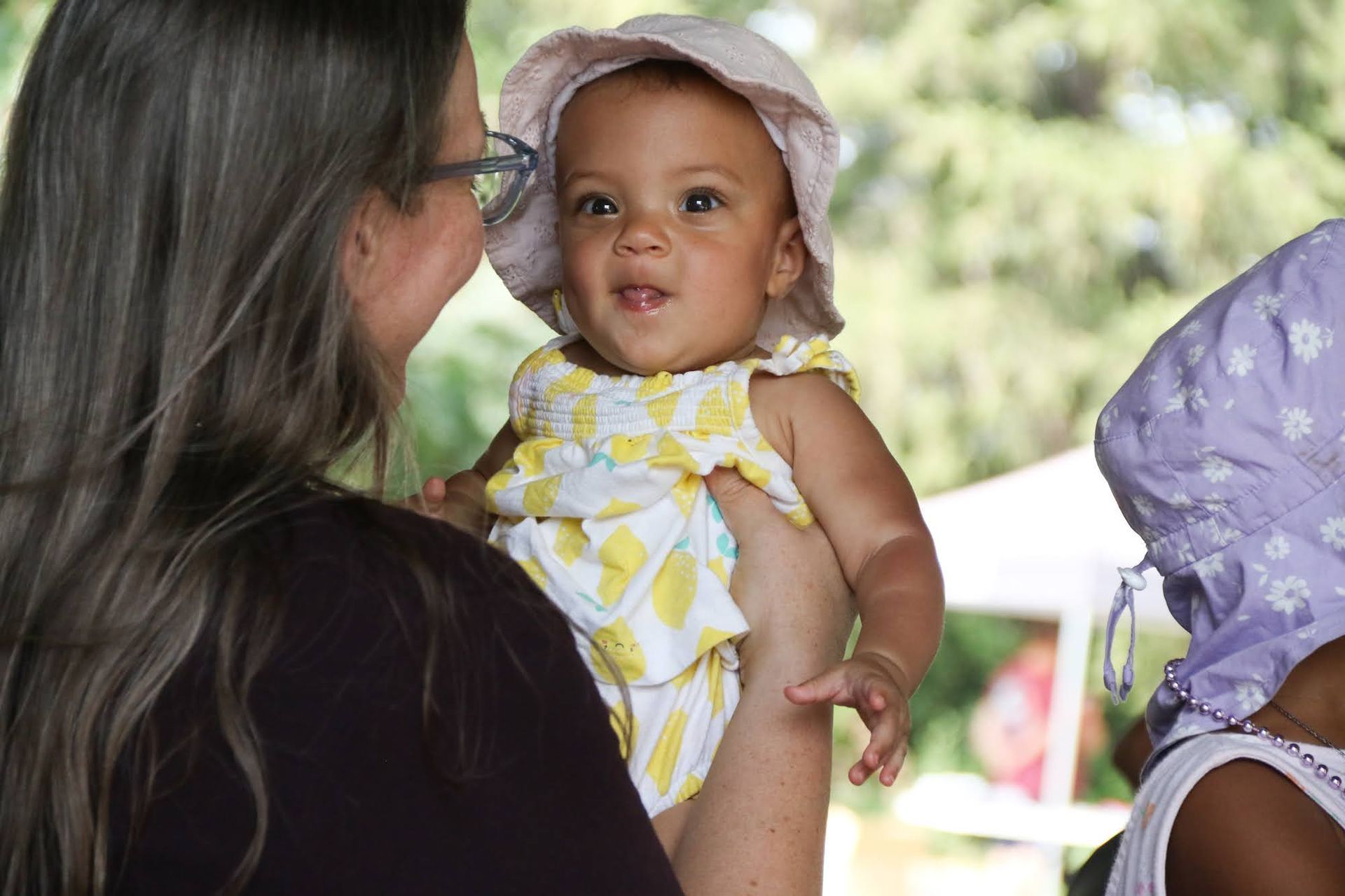 Woman holding a baby wearing a sun hat and yellow and white outfit; outdoor setting.