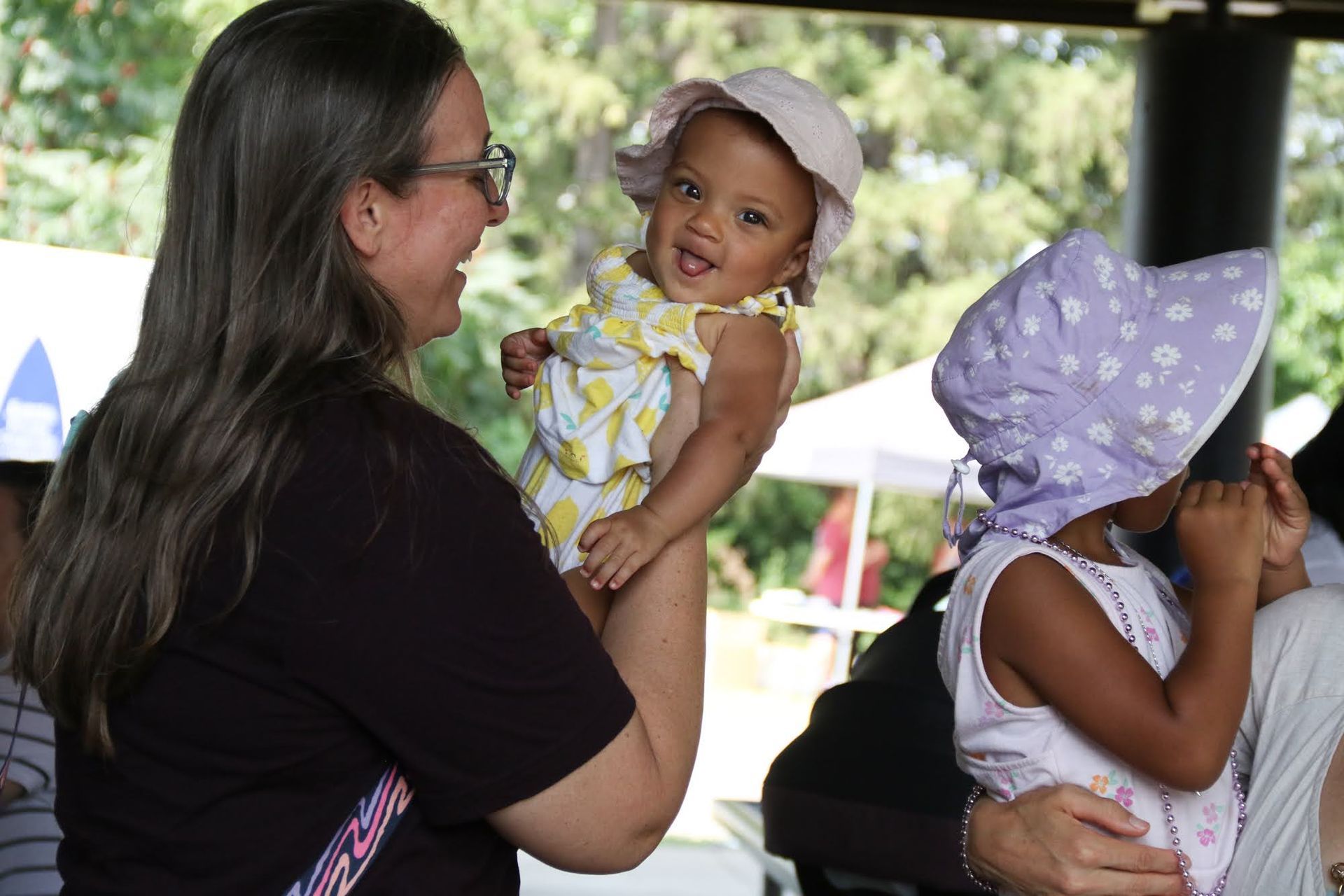 Woman holding a smiling baby wearing a sun hat outdoors, another child nearby.