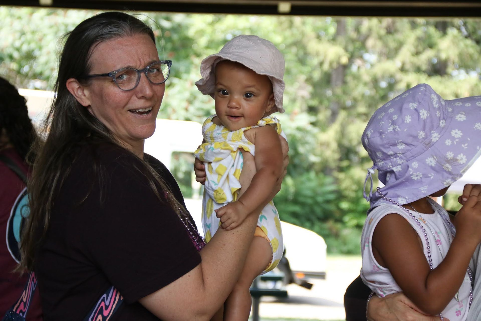 Woman holding baby wearing a sun hat, another child wearing a sun hat nearby, outdoors.