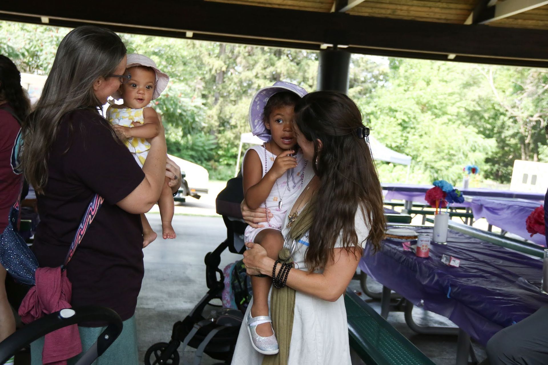 Two adults hold children at a park, near a decorated table.