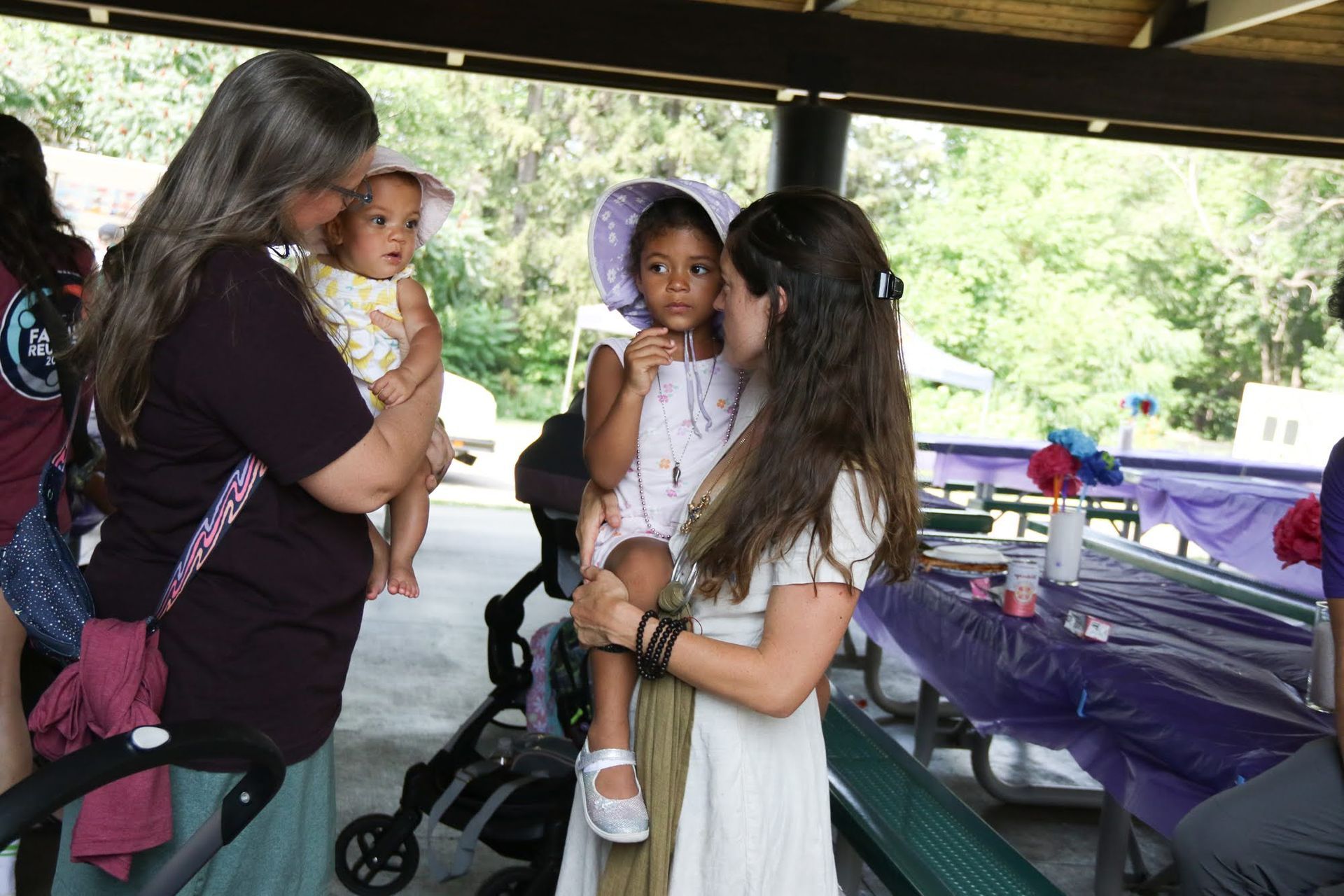 Two women holding children at a picnic table, other people in background.