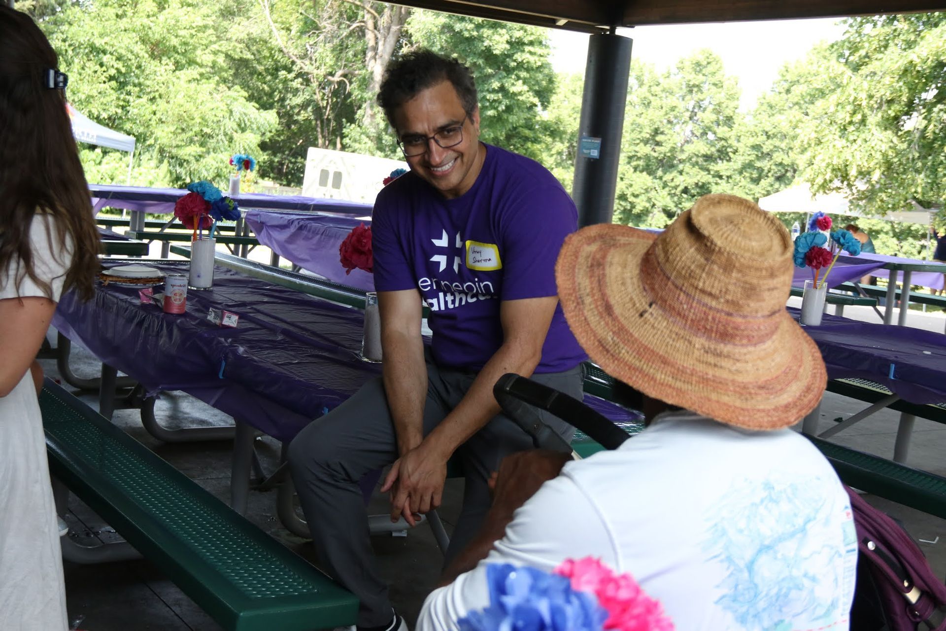 Man in purple shirt smiles while talking to a person wearing a straw hat at an outdoor picnic.
