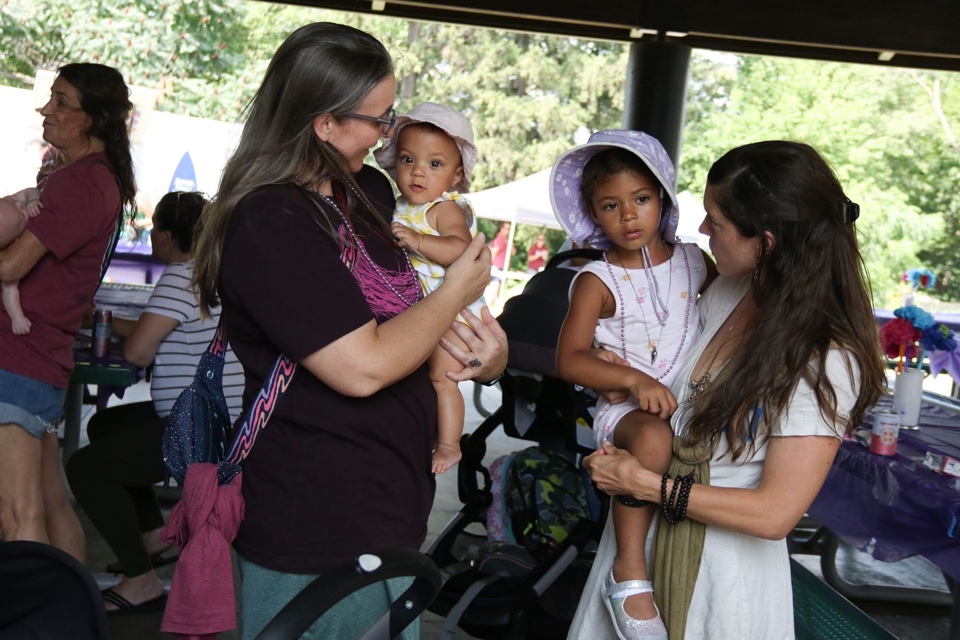 Two women holding children, smiling, at an outdoor gathering, other people visible in background.