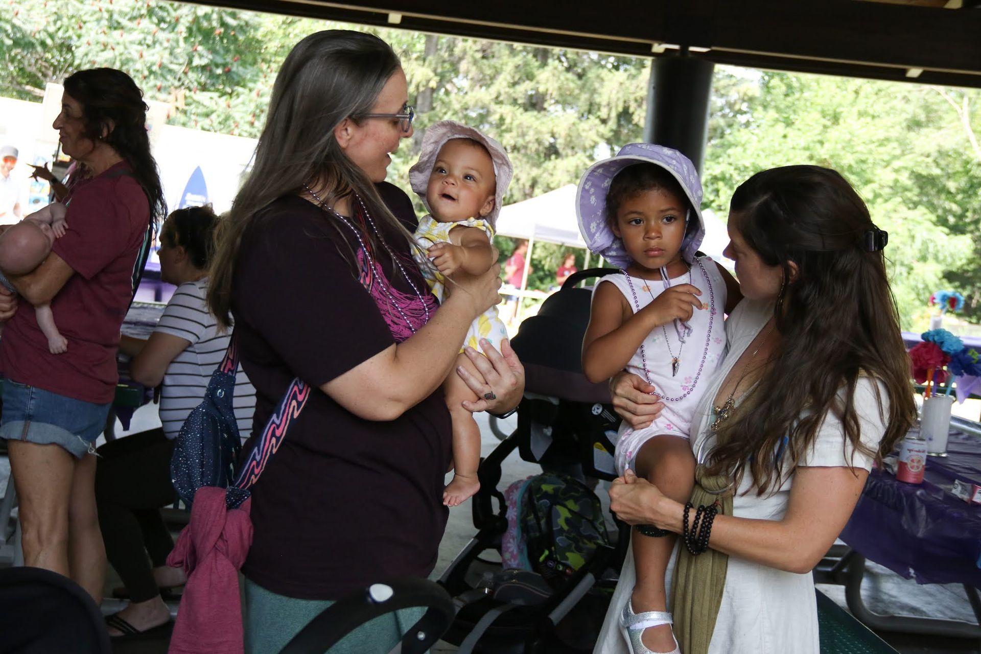 Two women holding toddlers at an outdoor event. One child wears a sun hat. People and tables are in the background.