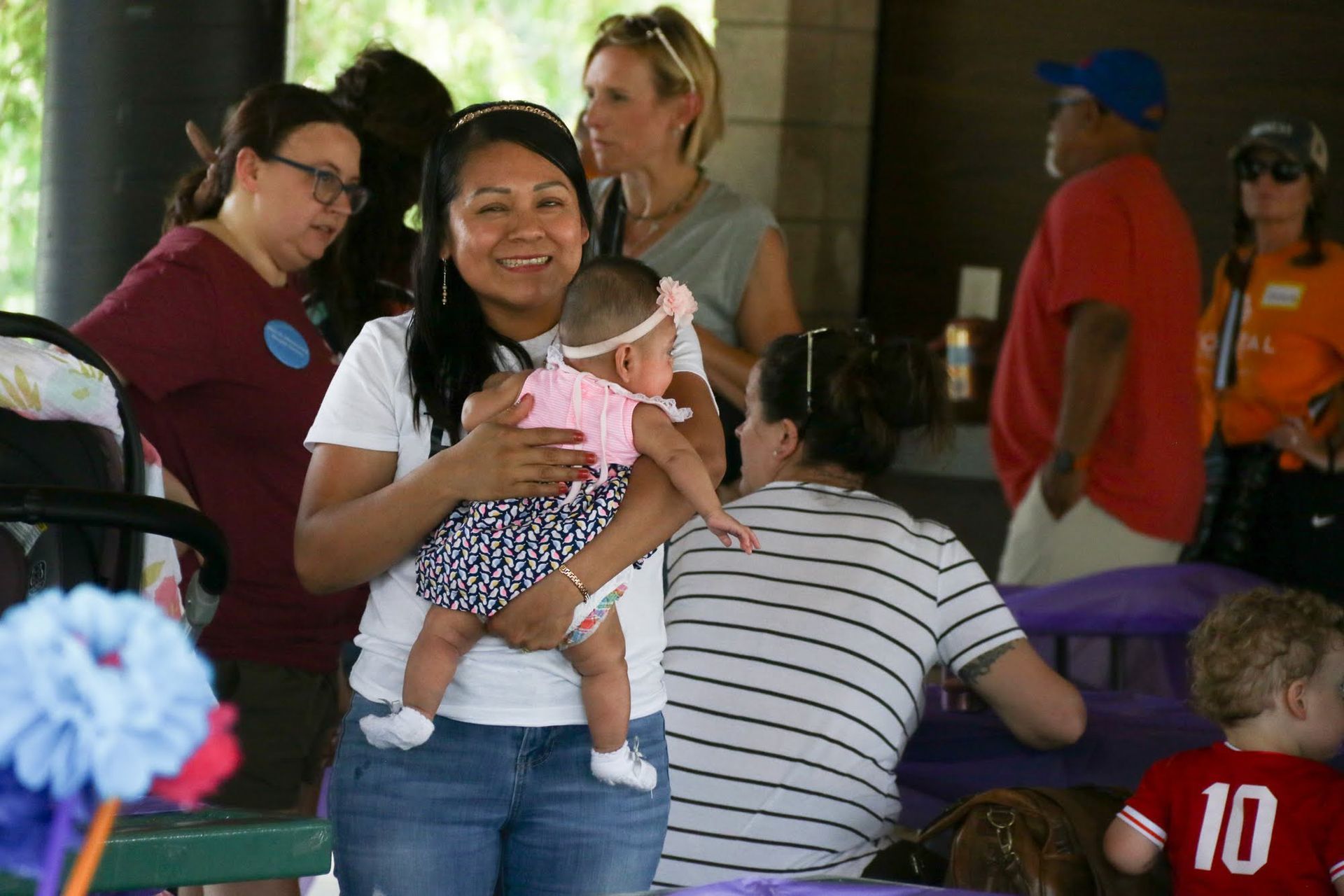 Woman holding a baby smiles at the camera at an outdoor gathering.