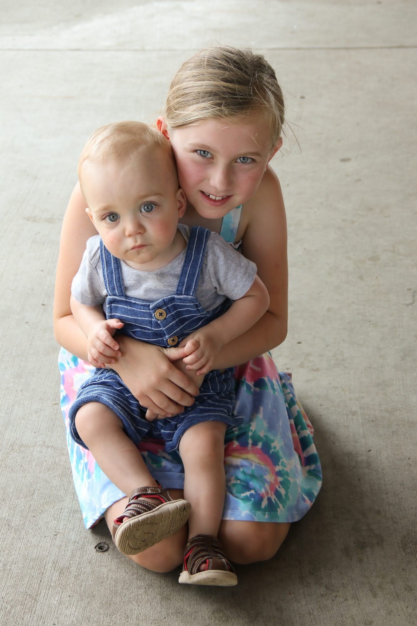 Girl kneeling, holding baby. They are both looking at the camera. Outdoor, concrete background.
