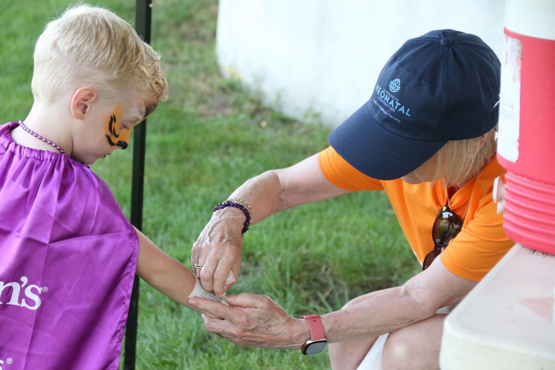 Woman putting a band-aid on a child's arm outdoors. Child has face paint and a purple cape.
