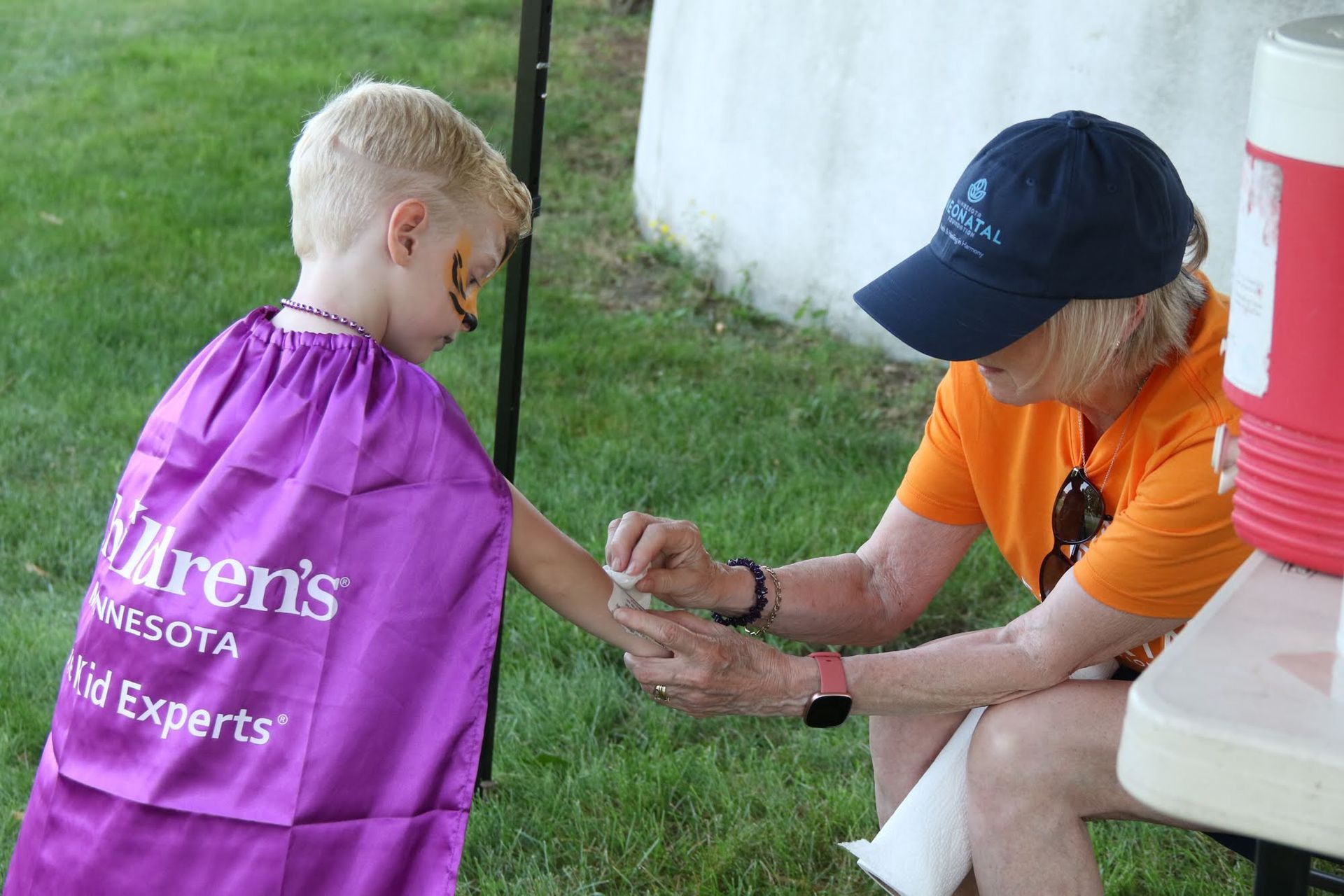 A person applies face paint to a child's arm outdoors; child wears purple cape.