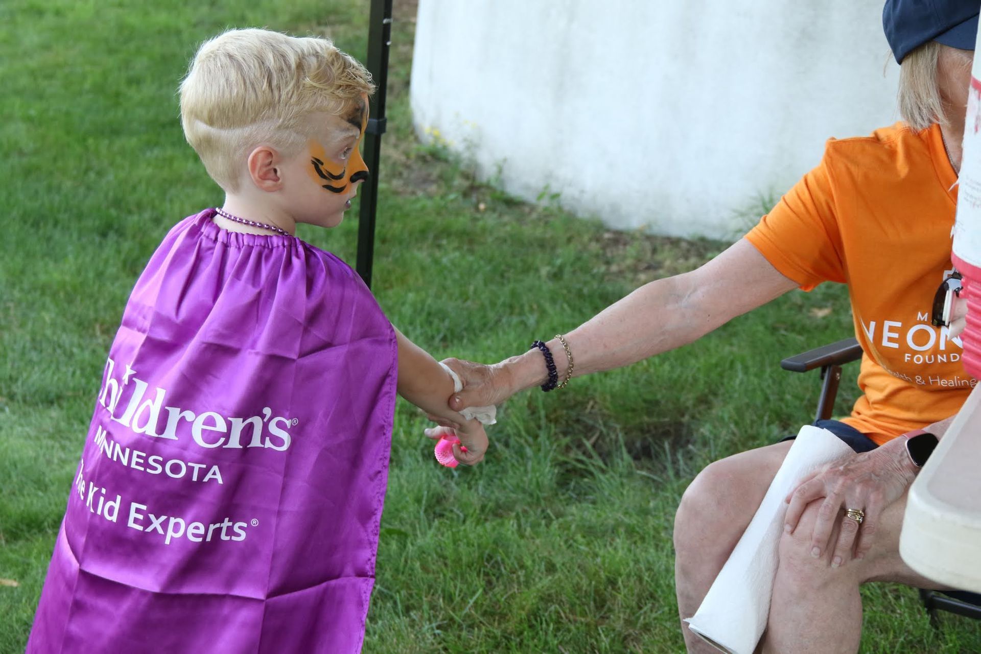 Child in purple cape with face paint shaking hands with person in orange shirt.