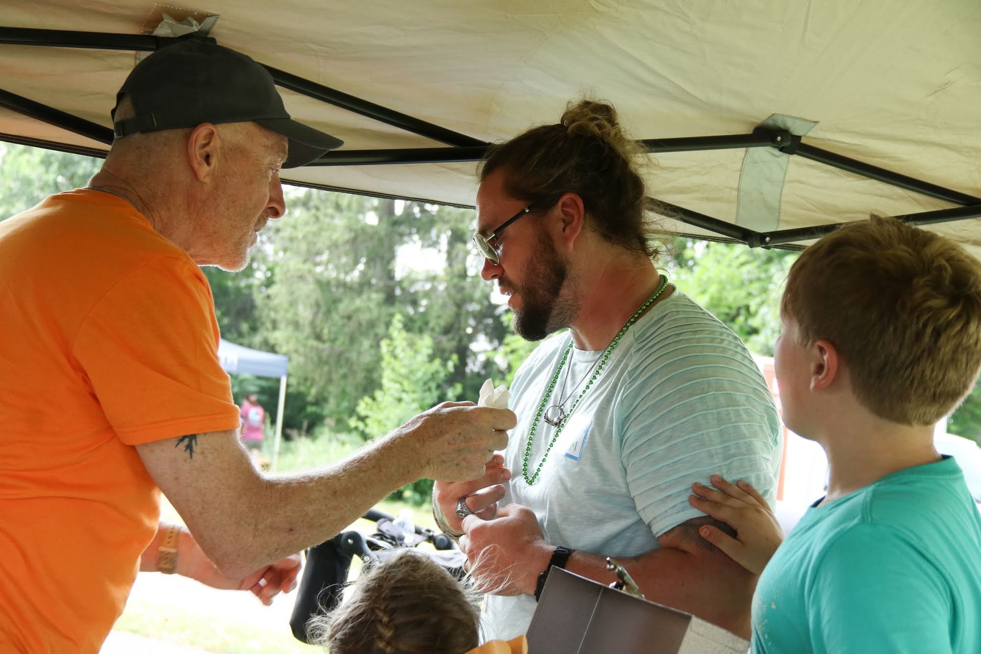 Man in orange shirt hands object to man with bun, child watches. Under a canopy, outdoors.