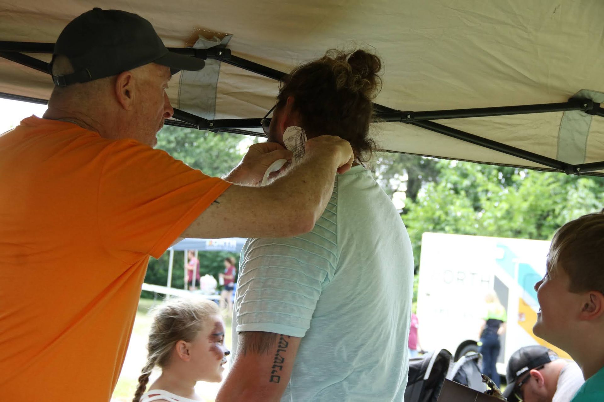 Man applying something to another's neck under a tent. Child watches, another with face paint in foreground.