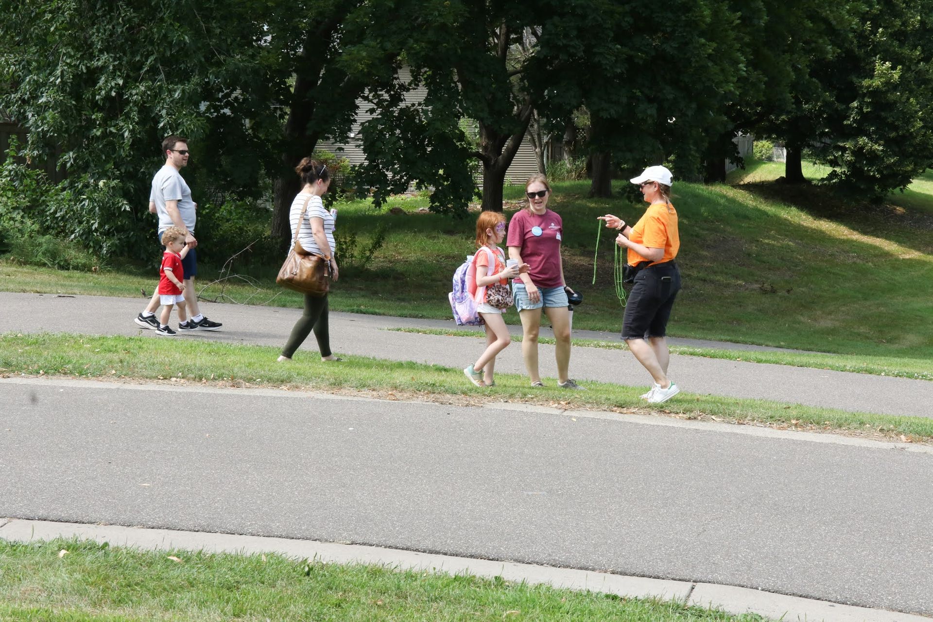 People on a paved path in a park. One woman gestures, talking to two young girls. Others walk in background.