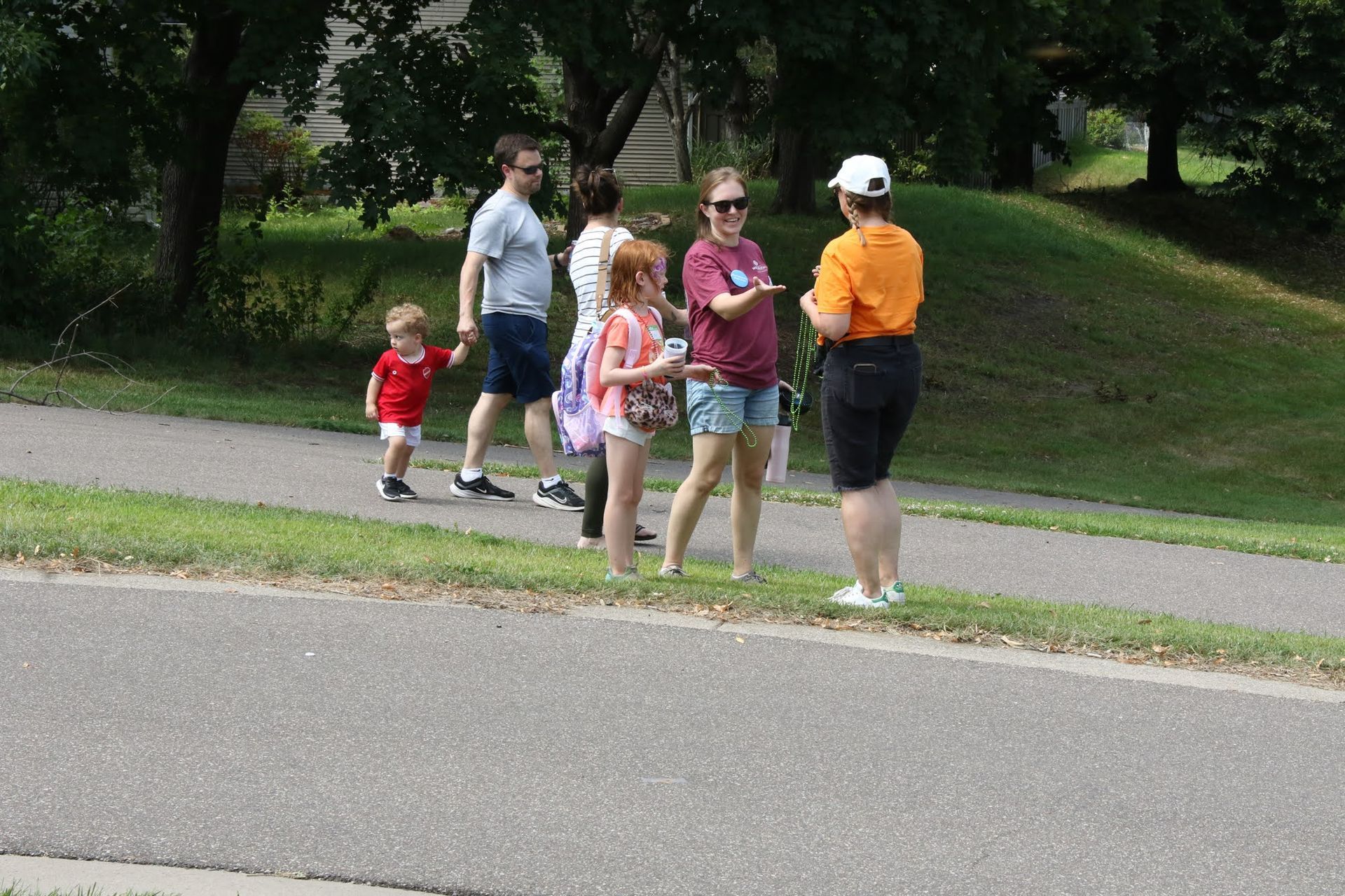 People on a paved path. A person in an orange shirt talks to a group holding bags. A grassy hill is in the background.