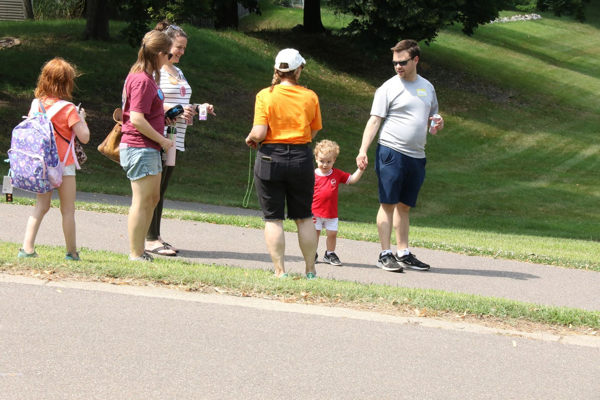 People walking on a paved path in a park. Two adults hold a young child's hand. Green hill in background.