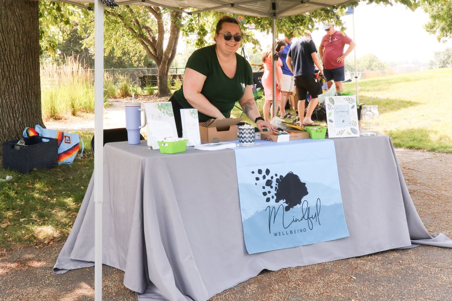 Woman at outdoor market stall; gray tablecloth, sign, customers in background.