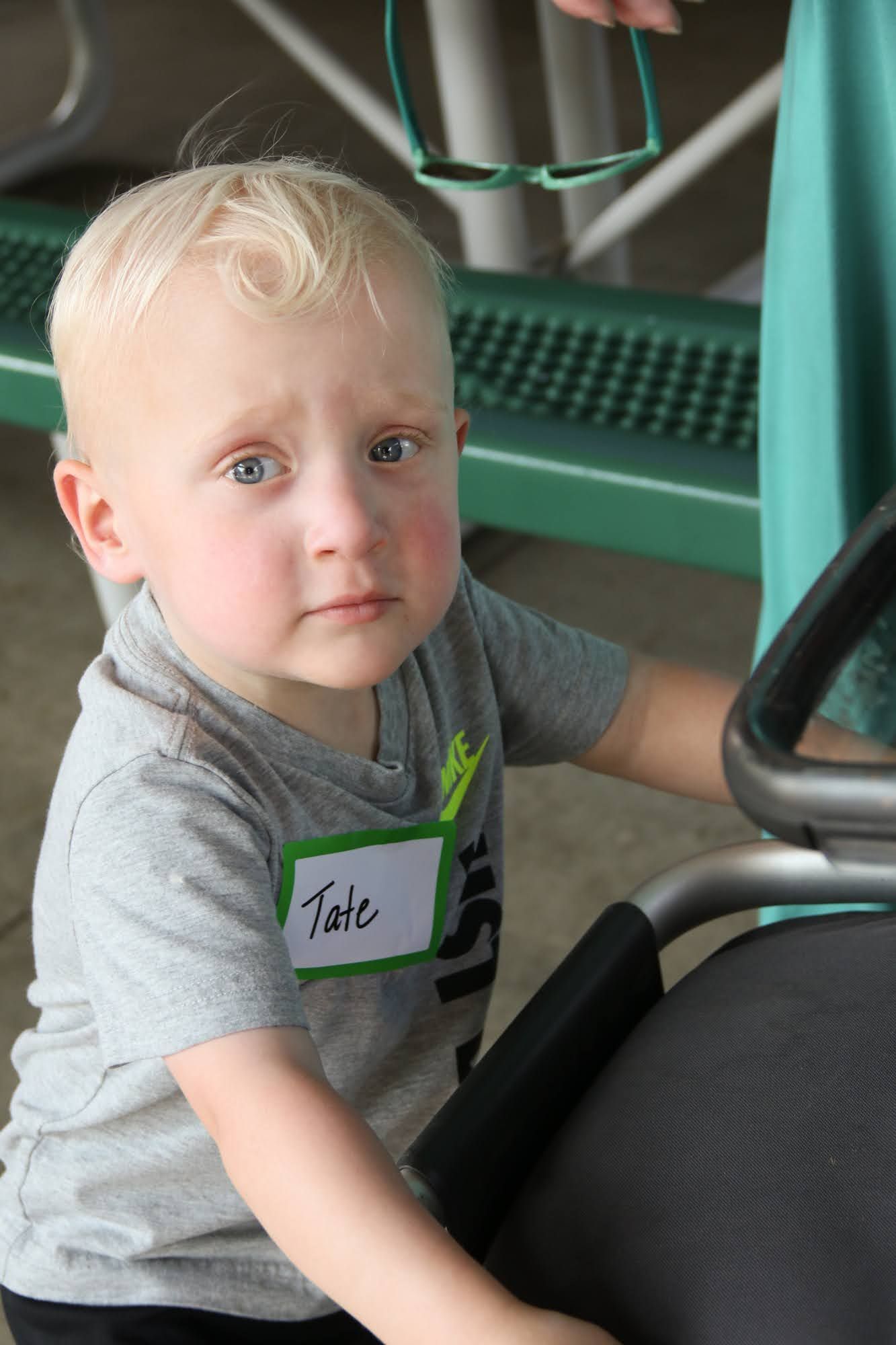 Blond toddler with a name tag, looking at the camera. He's near a chair, wearing a gray shirt, in an outdoor setting.
