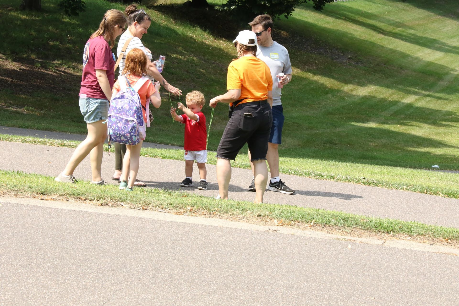 People standing on a paved path in a park. A small child reaches for someone's hands. Sunlight.