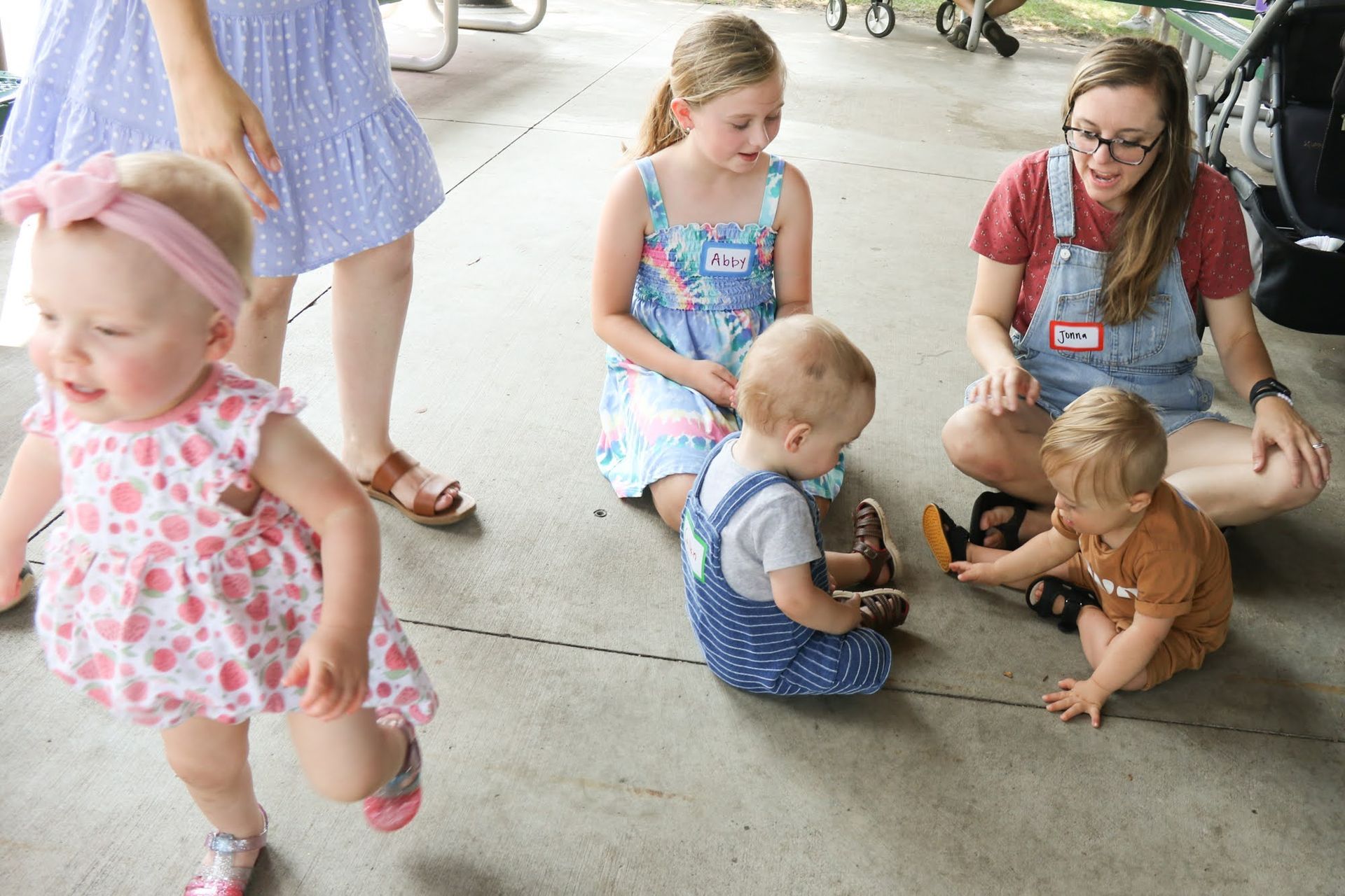 Children and a woman at a picnic. One baby runs. Others sit, looking at something. Overalls, dresses.