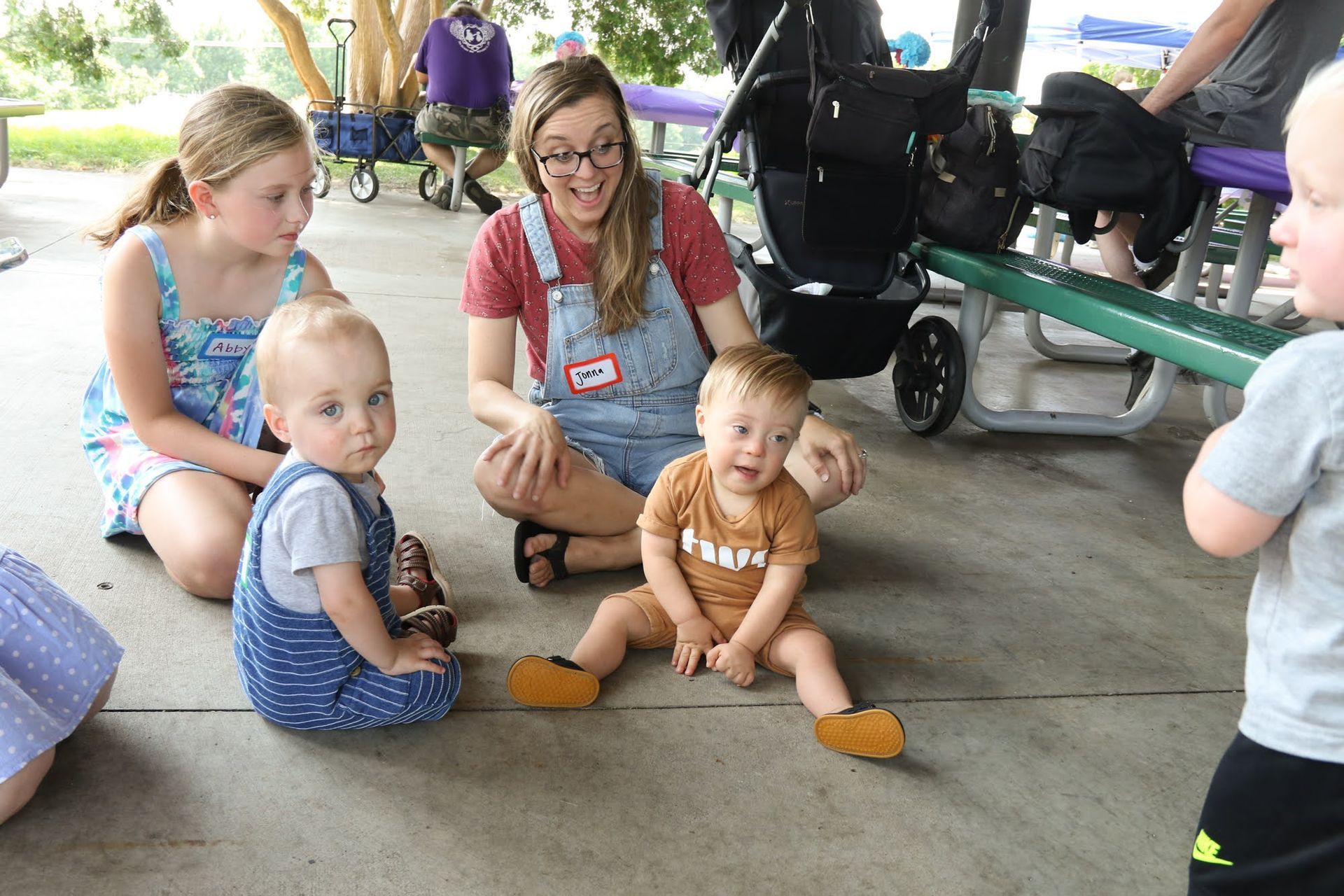 A woman sits with three babies and a child in an outdoor setting. The babies are smiling and looking at the woman.
