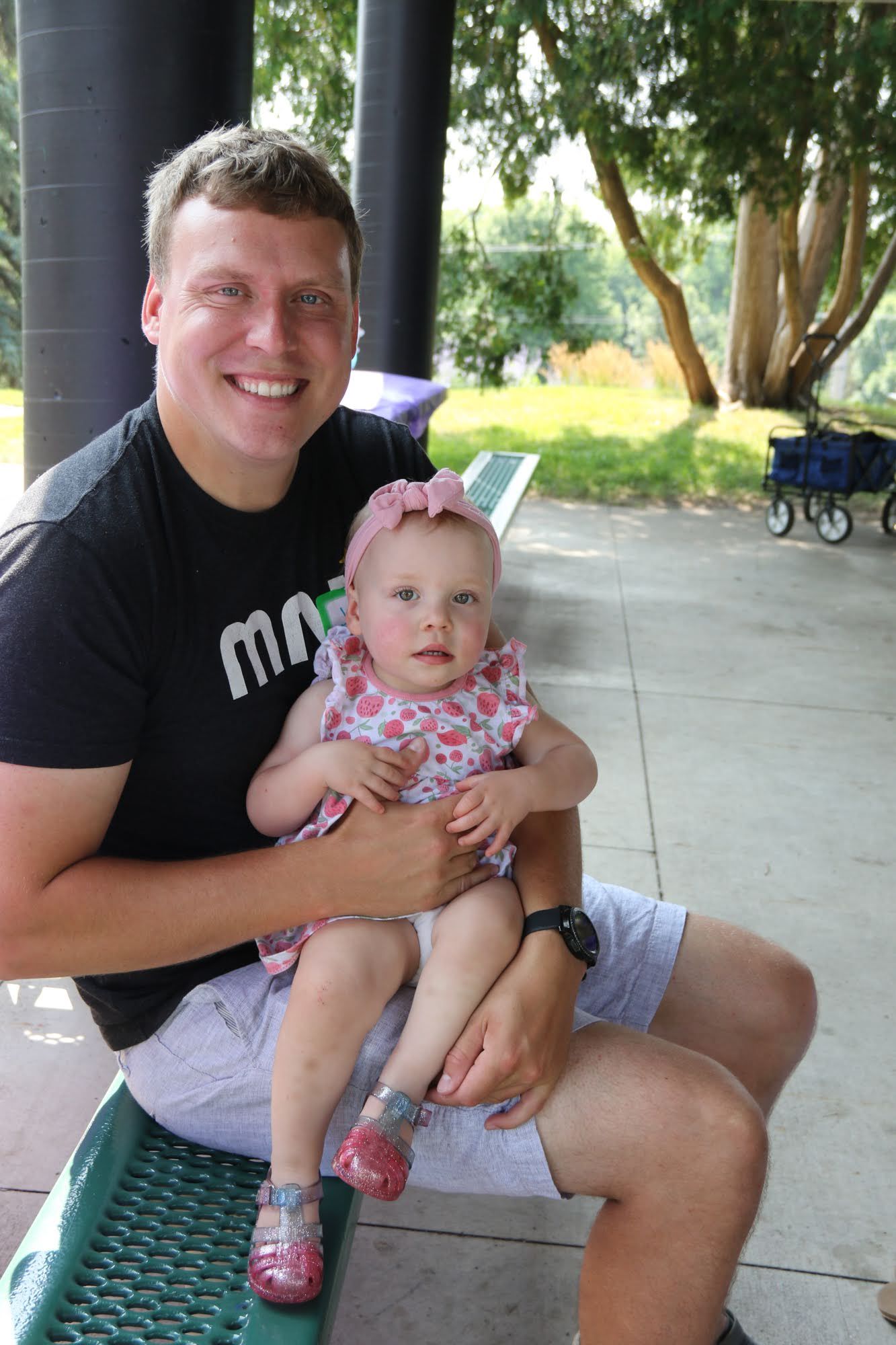 Man seated, holding a toddler wearing a pink floral dress, outside. Both smiling.