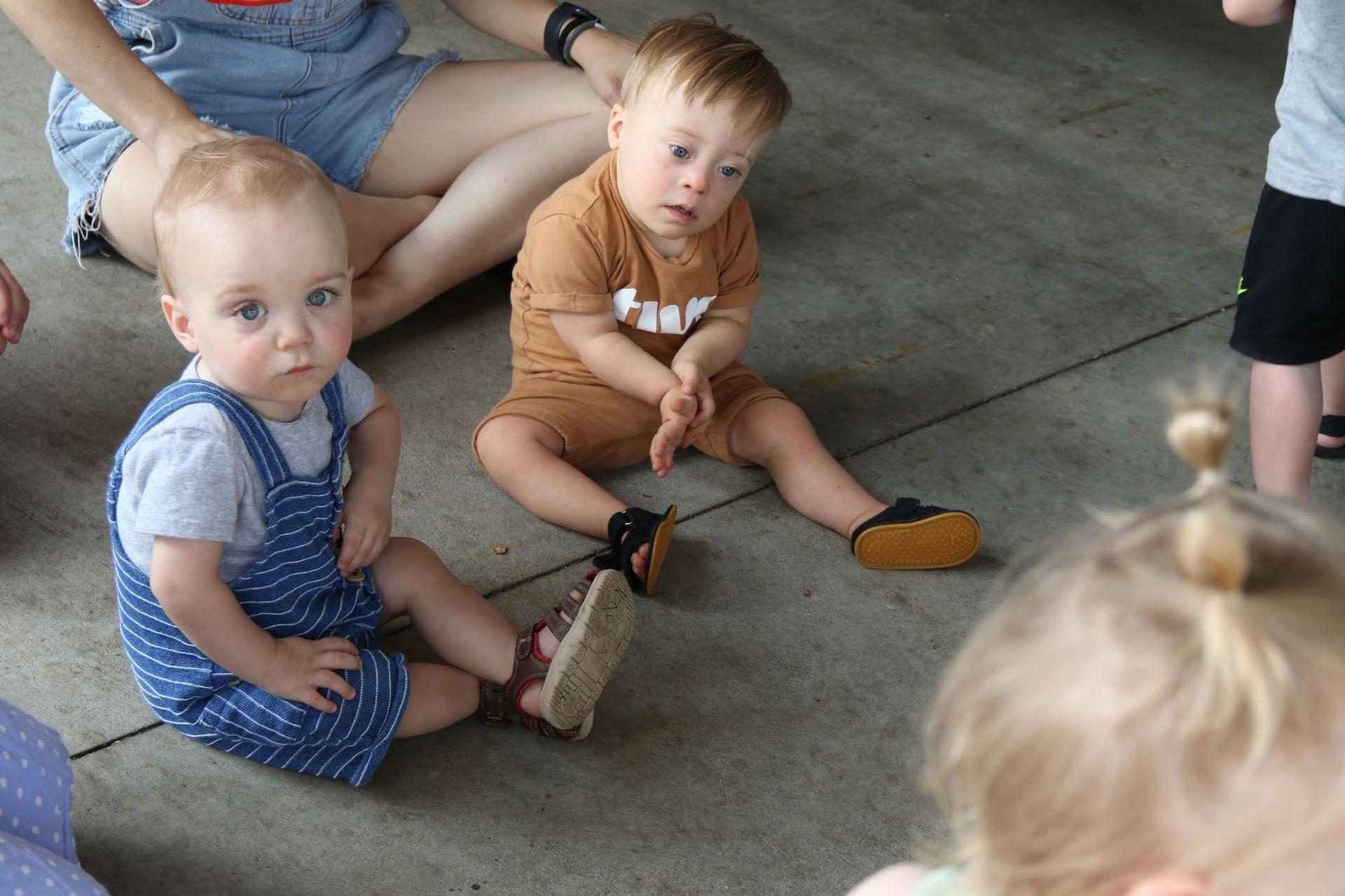Two toddlers and a person with blue shorts sit on the floor, looking forward.