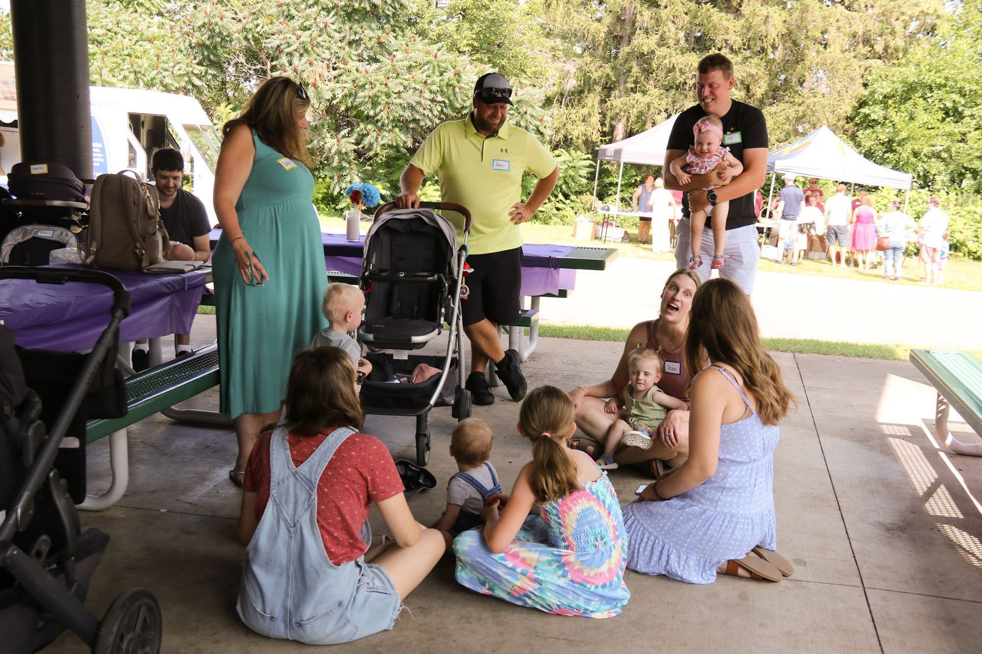 People gathered at a park shelter. Babies, adults, strollers, and picnic tables are present.