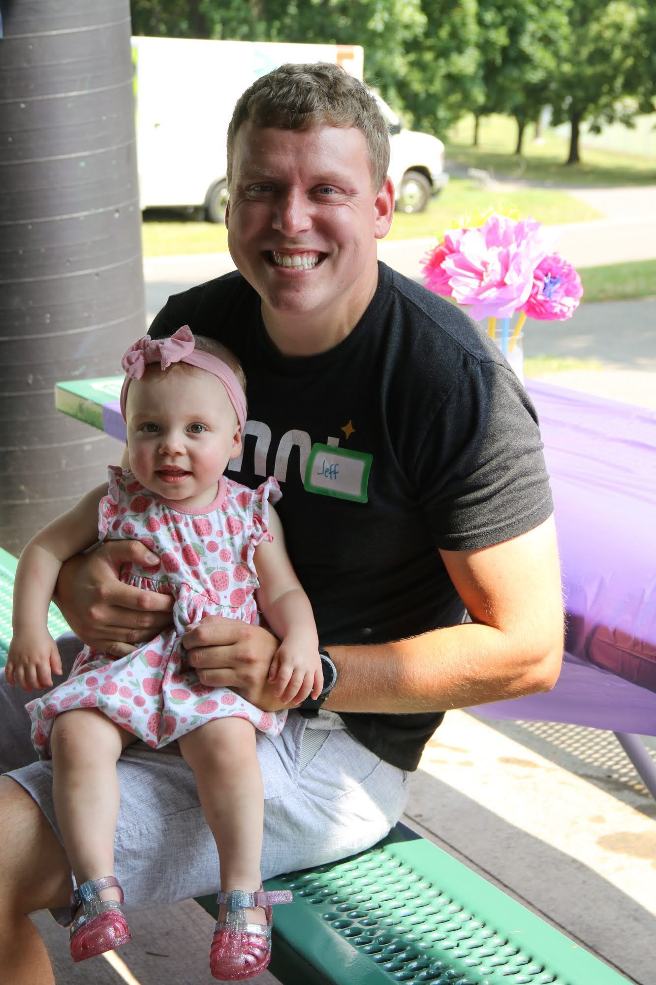 Man smiling, holding baby girl wearing pink dress, sitting at a picnic table.