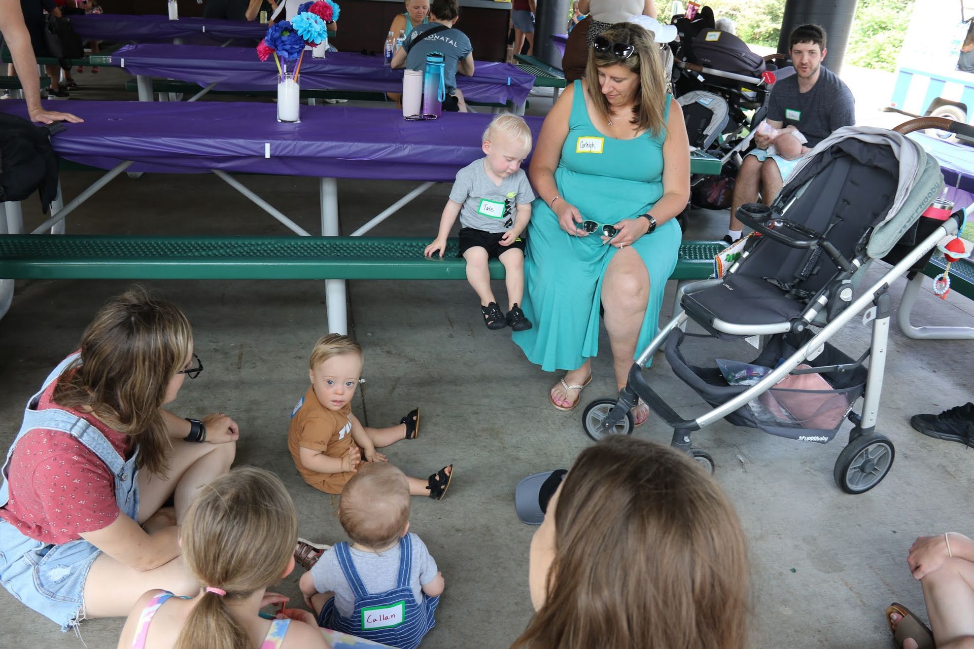 Group of people with young children in a park. Woman in teal dress, stroller, kids sitting and playing.