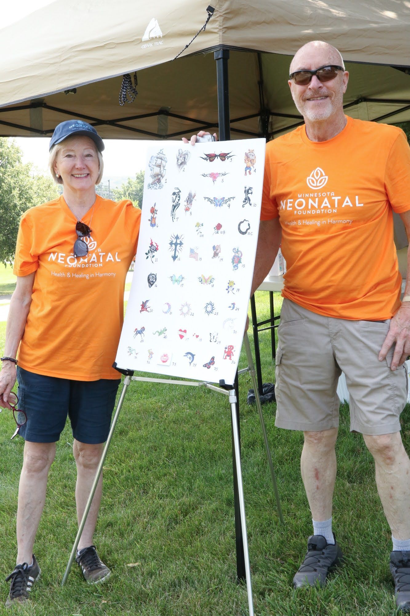 Two people in orange shirts hold a poster of symbols outdoors, under a tent.