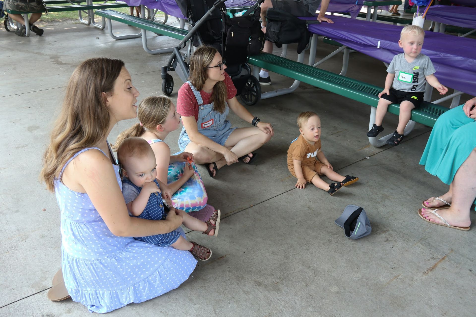 People and children sit in a park, talking. A baby is held, while others sit nearby.