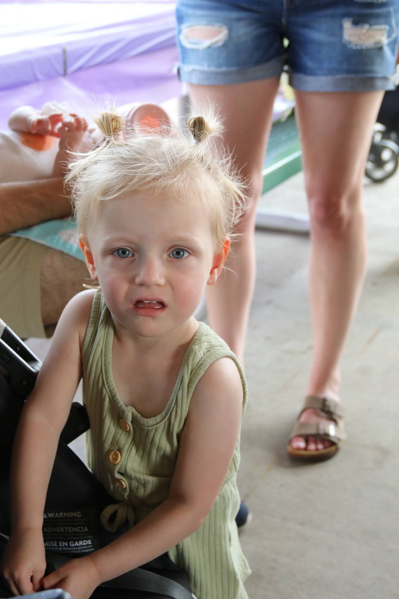 Blond toddler with messy buns looks at the camera, wearing a green dress. A person stands behind her in jean shorts and sandals.