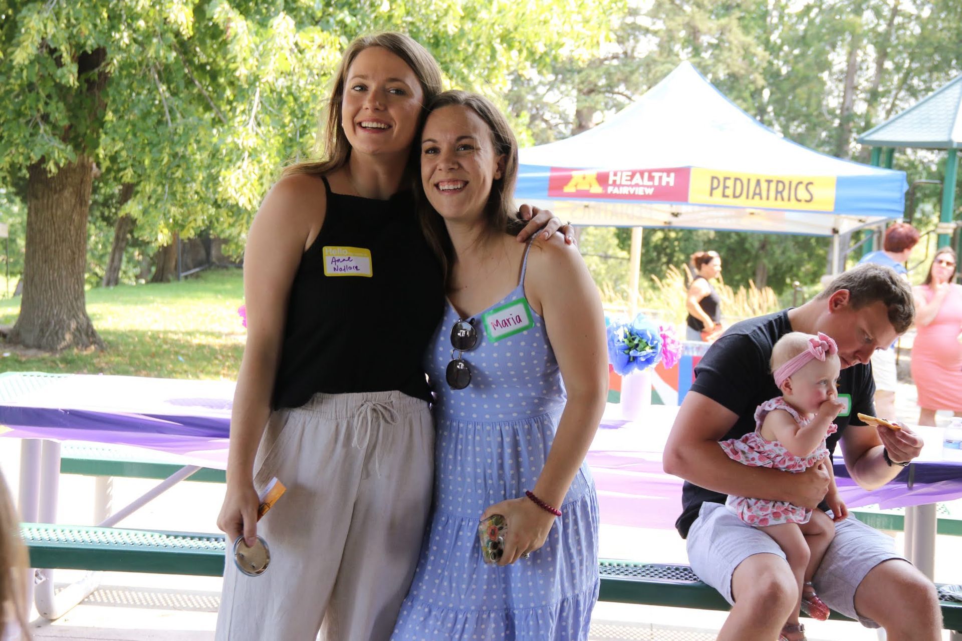 Two women with name tags pose smiling outdoors. A man holds a baby. Event tent in the background.