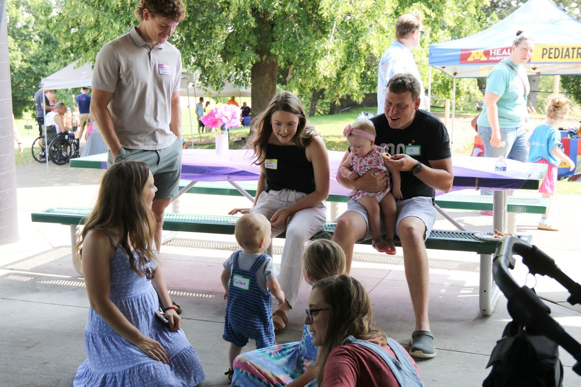 People at a park picnic: babies, young adults, seated at a table. Tents and trees in the background.