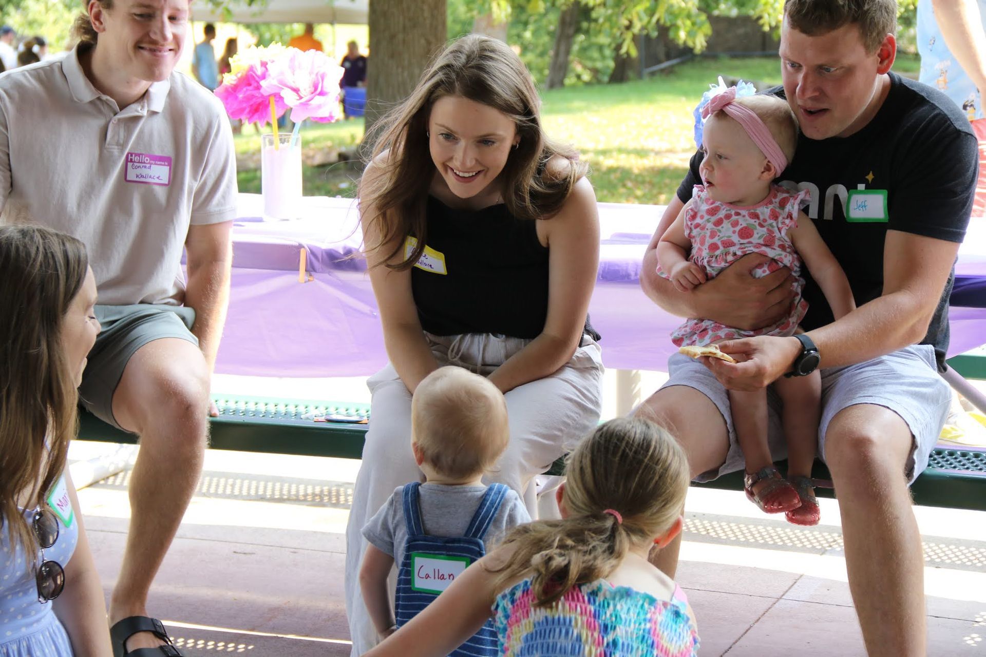 People at an outdoor gathering, interacting with children. Woman smiles at a toddler; man holds a baby.