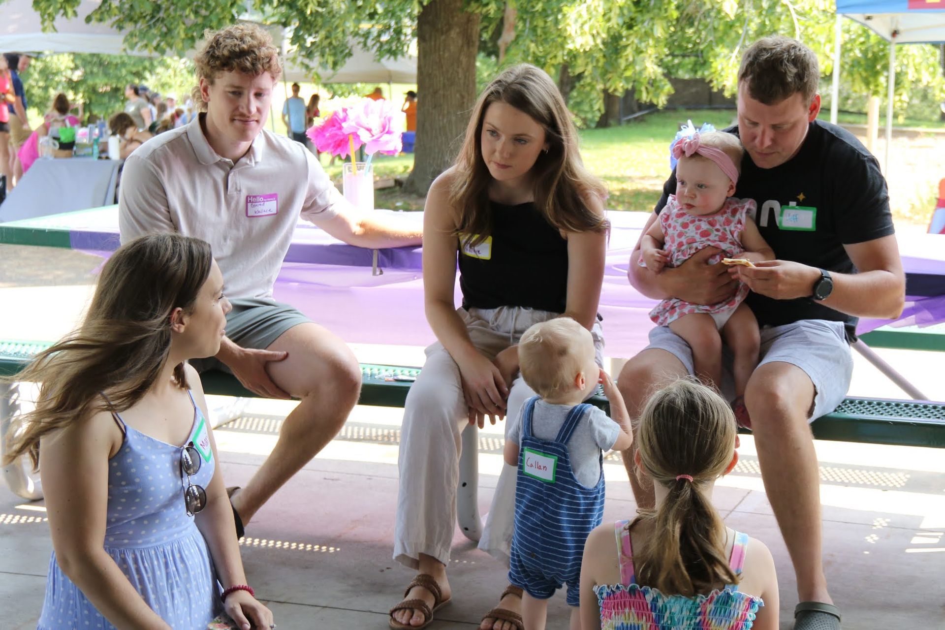 Group of people outdoors with children; some are seated, interacting near a stage.
