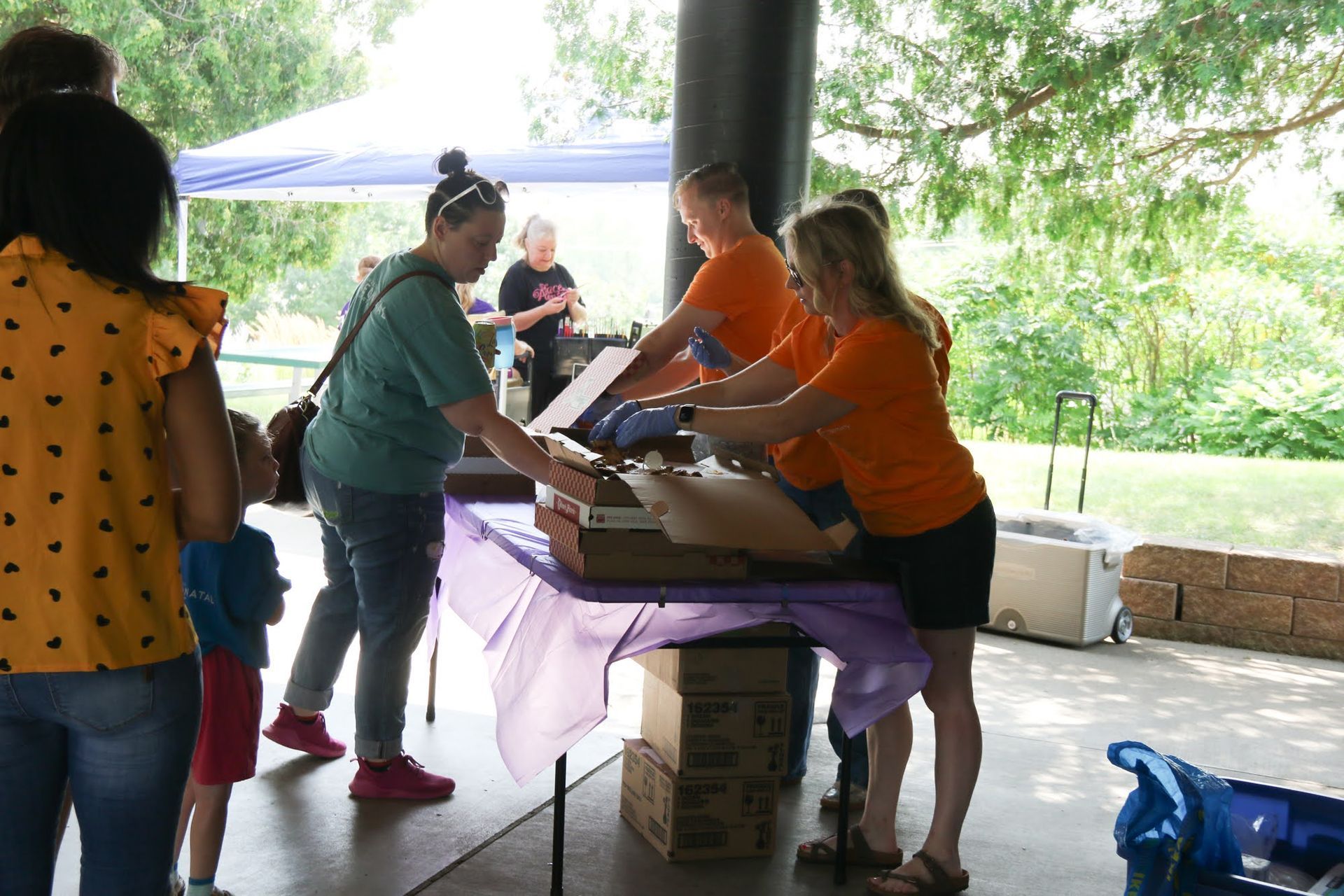 People serving food at an outdoor event under a shelter. Two wear orange shirts and assist others at a table.